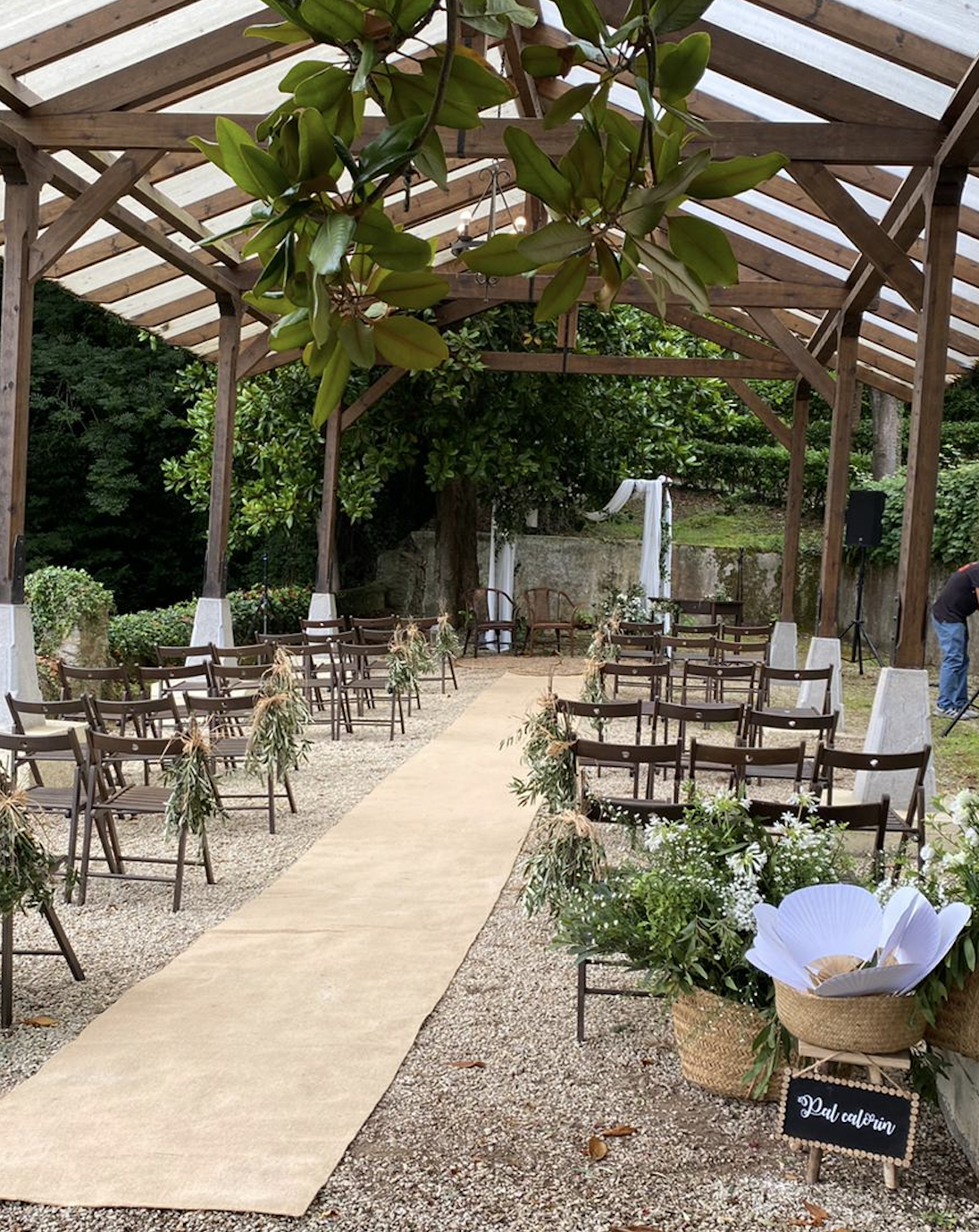 Escenario decorado para una ceremonia al aire libre con sillas de madera, camino de tela, plantas y flores, en un espacio techado con estructura de madera, y un altar con tela blanca en el fondo.