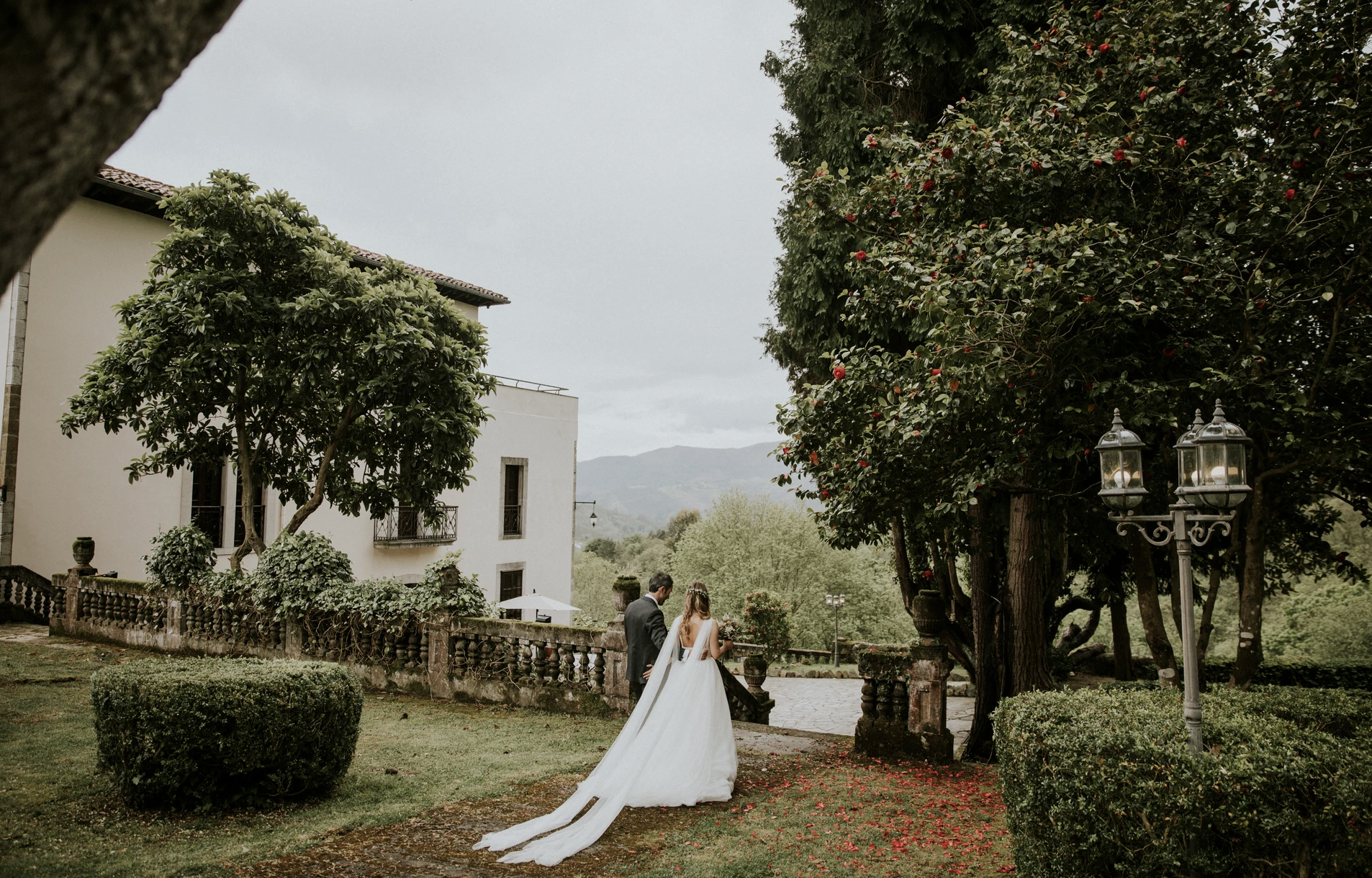 Pareja de novios caminando en un jardín con árboles y arbustos, durante el día, en un evento de boda.