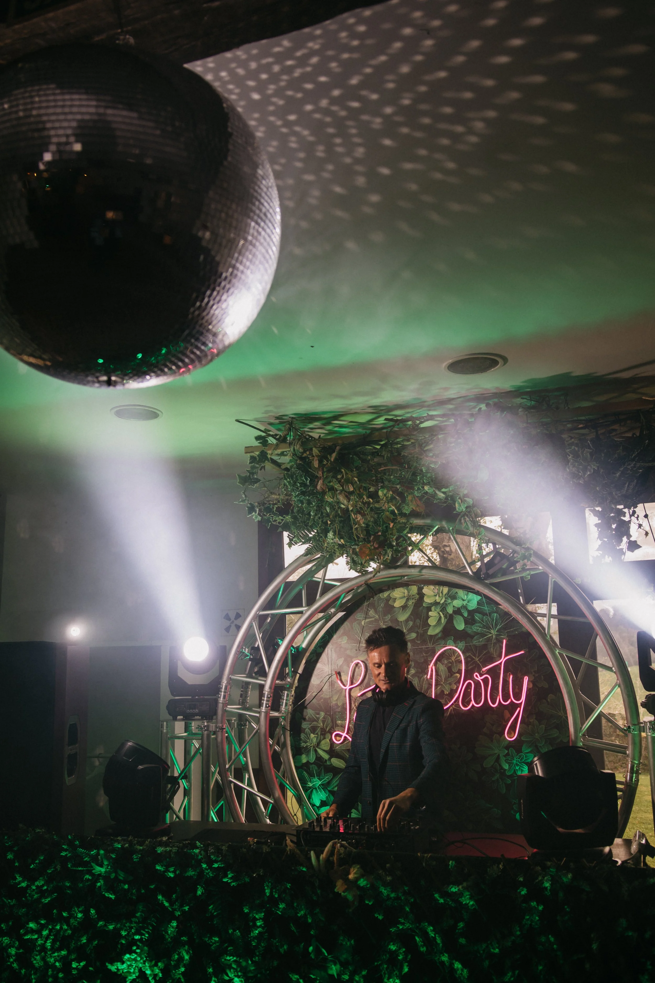 Un DJ mezclando música en una fiesta con decoración verde, un letrero de neón que dice 'Party' y una bola de espejos en el techo.