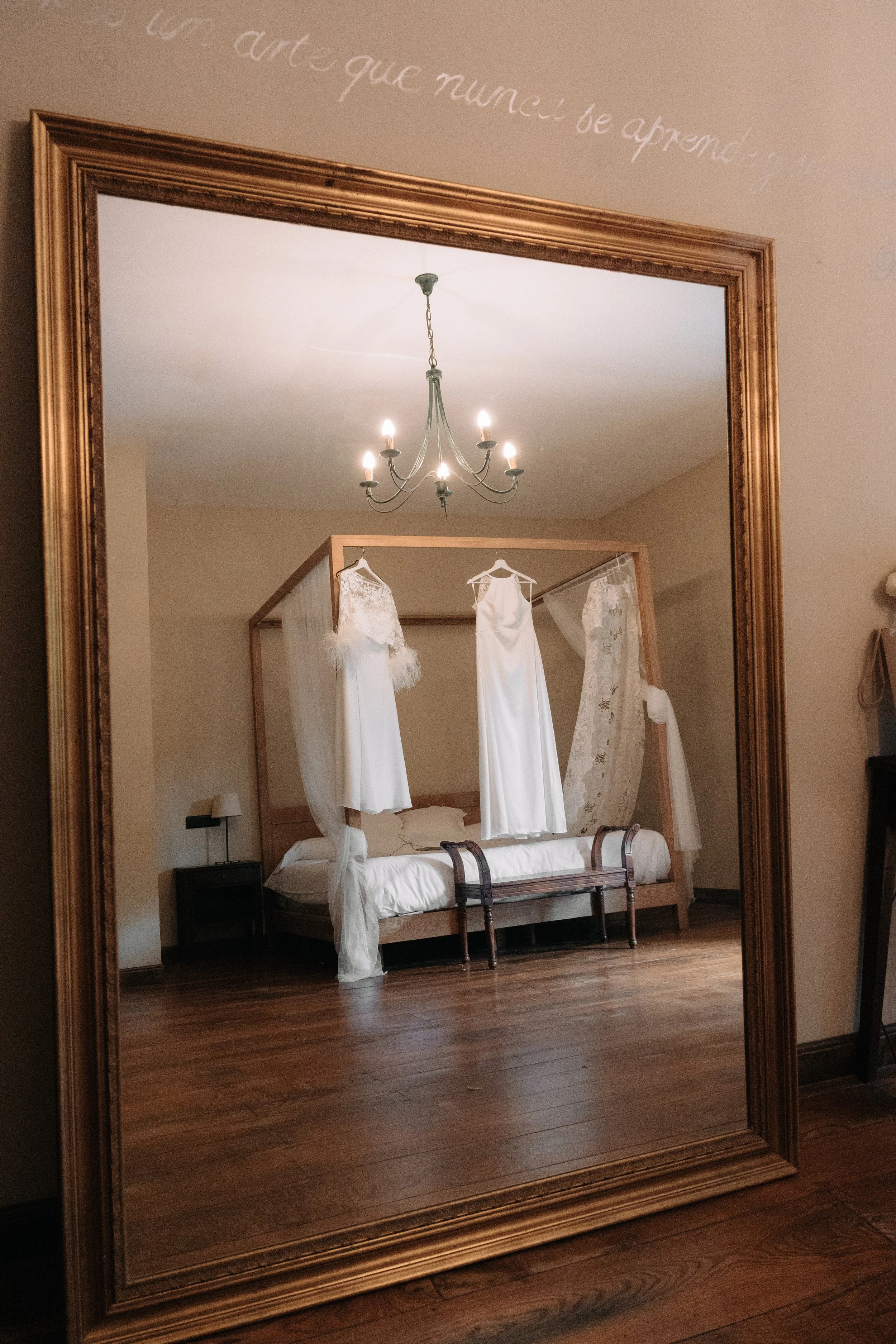 Espejo grande con marco dorado que refleja una habitación con camas y cortinas blancas, colgado un candelabro con cinco luces, y en la pared se puede leer una frase en cursiva en español.