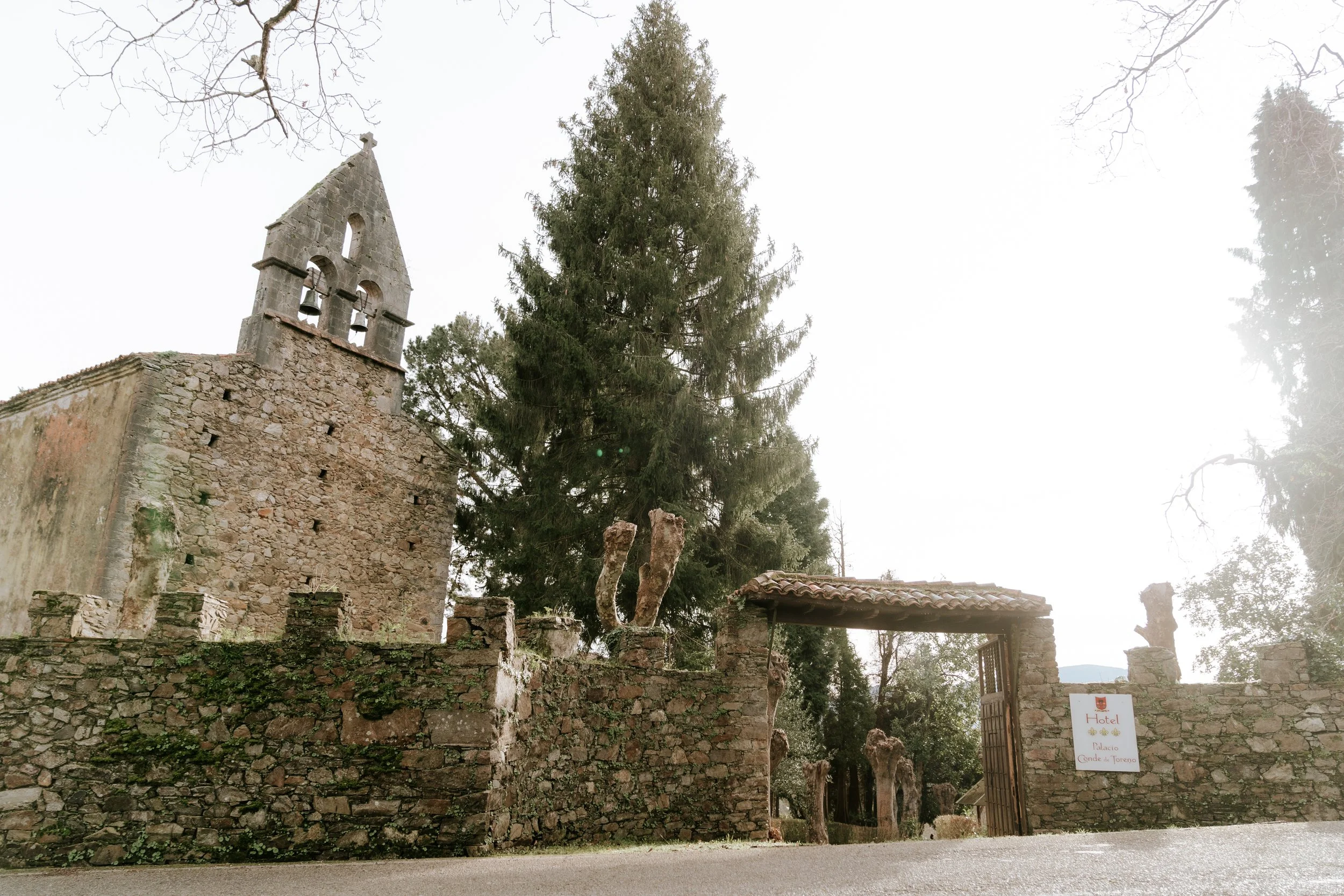 Antigua estructura de piedra con torre y campanario, árboles grandes alrededor, entrada con puerta de madera y cartel que dice 'Hotel Palacio Cande de Tereno'