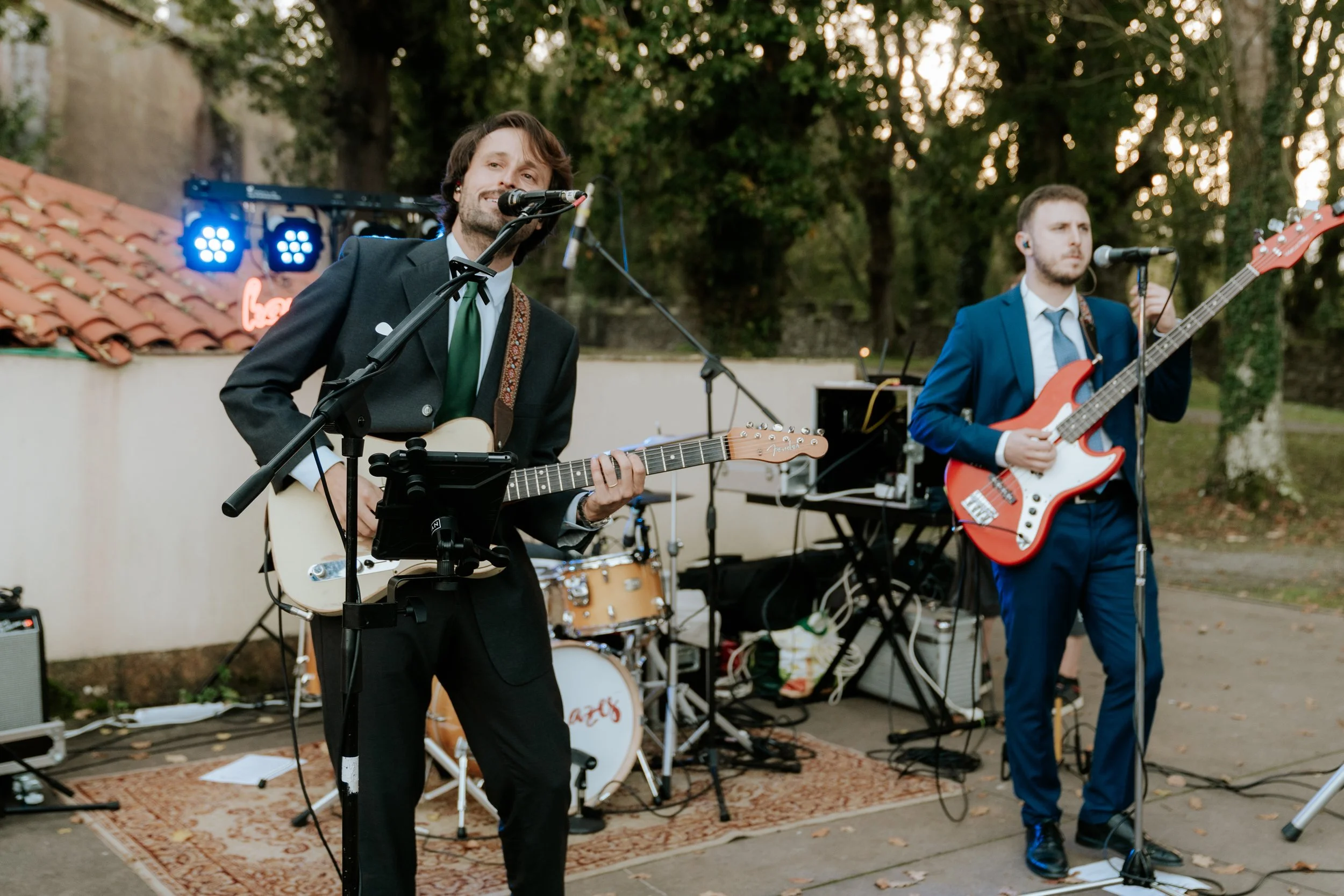 Grupo de músicos tocando en un evento al aire libre, con instrumentos como guitarra y bajo, en un ambiente de tarde con árboles de fondo.