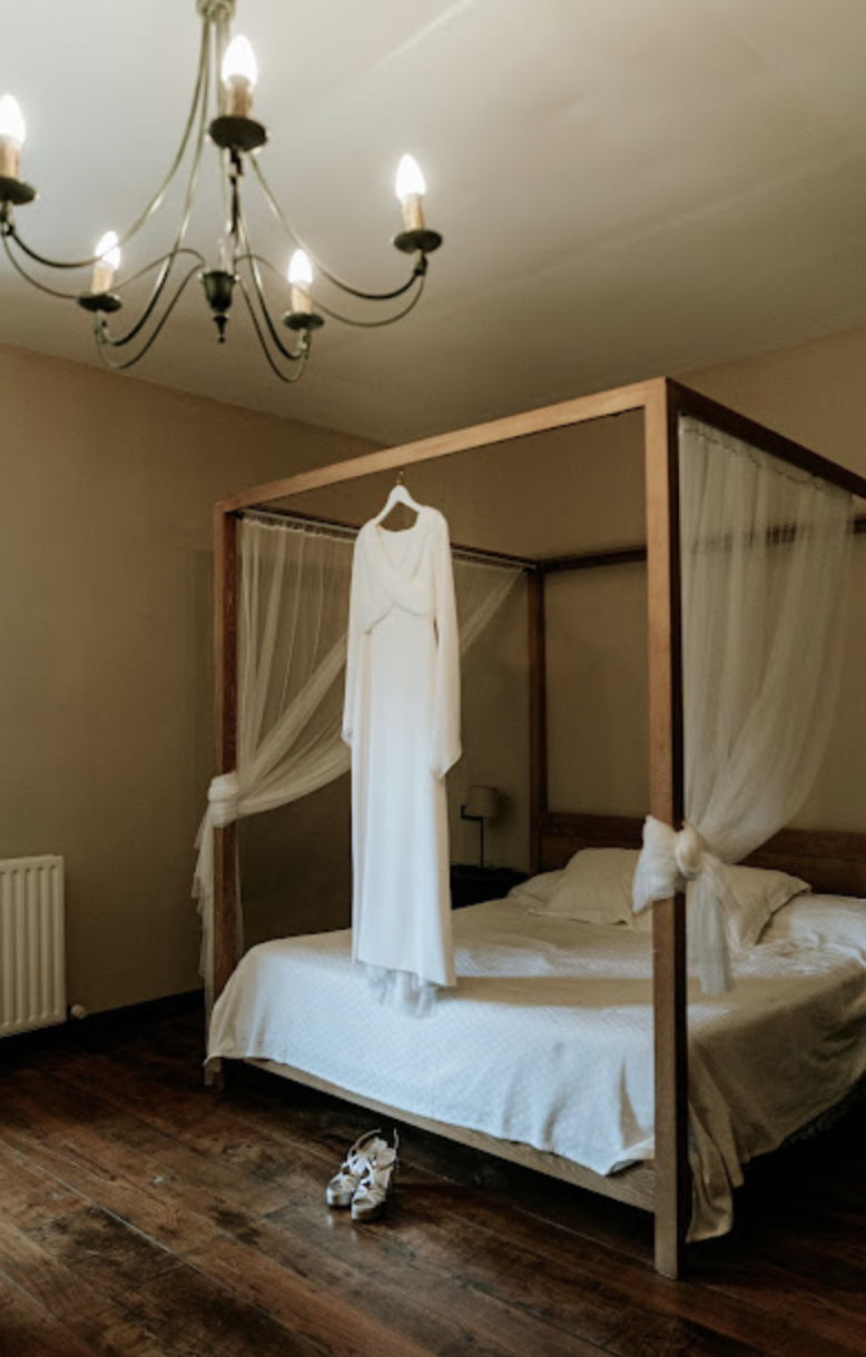 Habitación con cama de cuatro postes, con cortinas de encaje, y ropa de cama blanca. Un vestido blanco cuelga del poste del lado de la cabecera y un par de zapatillas blancas están en el piso junto a la cama. Un candelabro con varias velas ilumina la