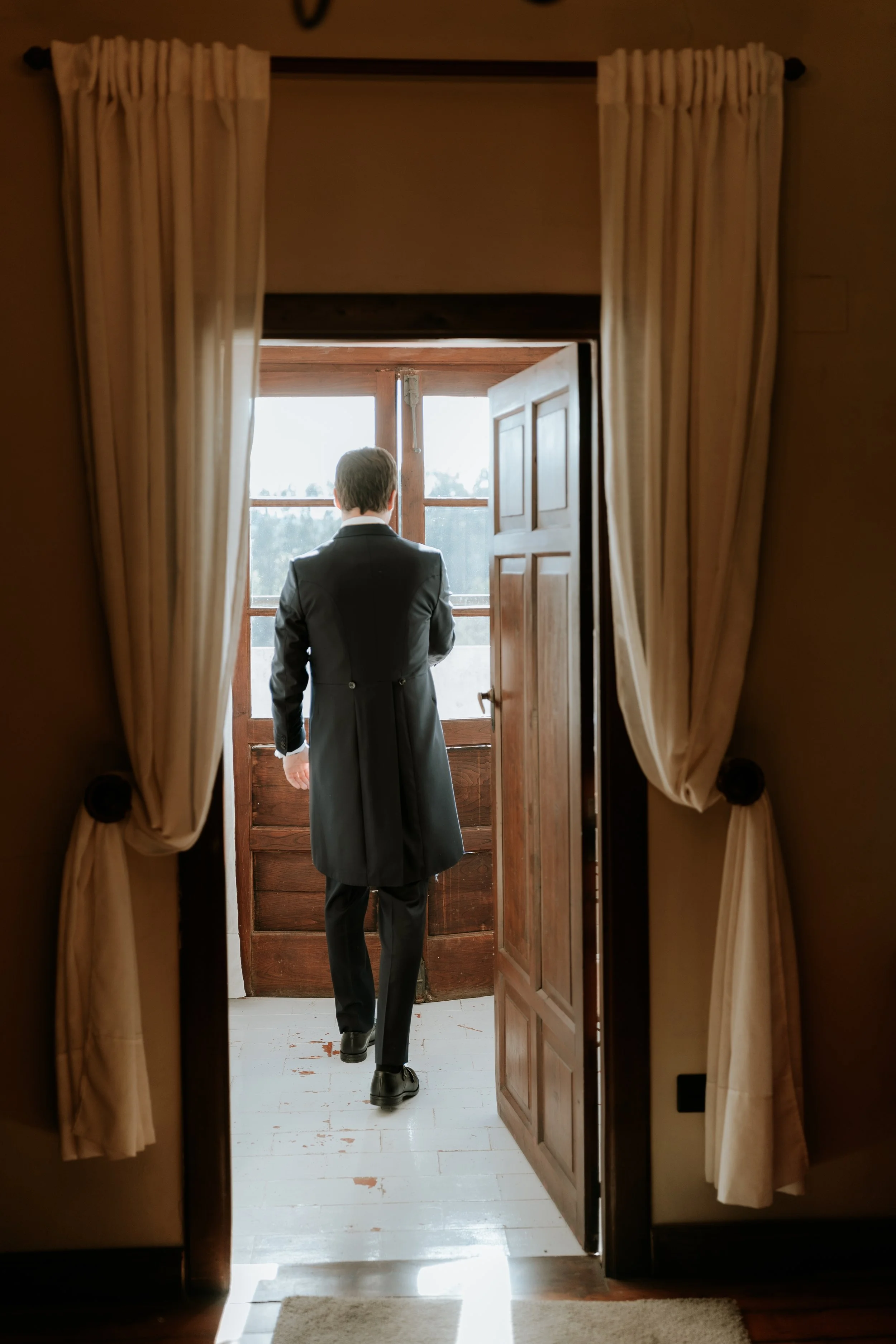 Hombre en traje negro de gala parado en una puerta de madera que da a un balcón o terraza exterior, con vista al horizonte y árboles en la distancia, visto desde dentro de una habitación con cortinas beige.