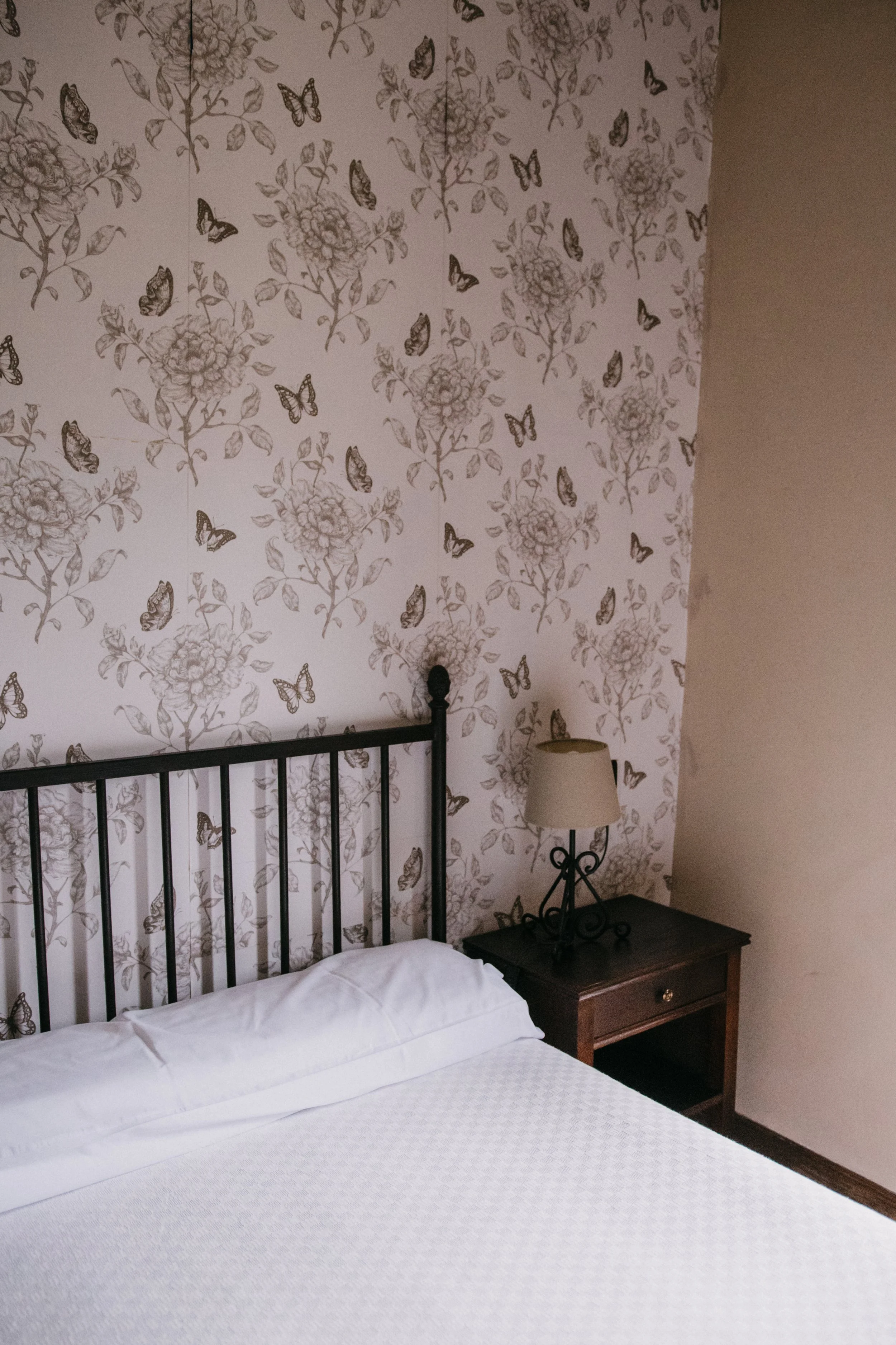 Habitación con pared decorada con papel tapiz que tiene flores y mariposas. Cama con sábanas blancas y cabecera de metal negro. Una lámpara de mesa de color beige en una mesita de madera a la derecha de la cama.