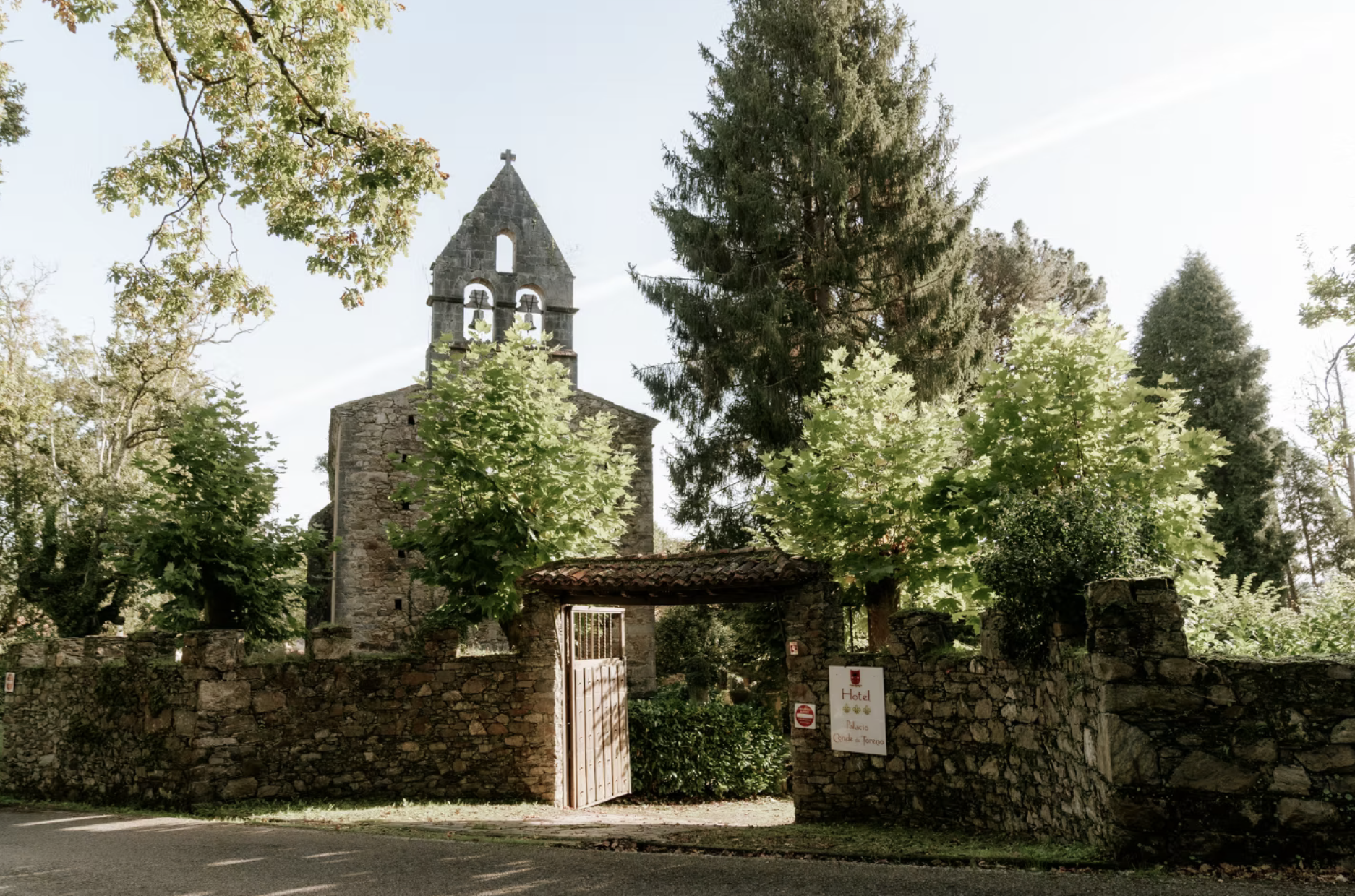 Una iglesia antigua construida con piedra rodeada de árboles grandes y una pared de piedra con una puerta de madera, en un día soleado.