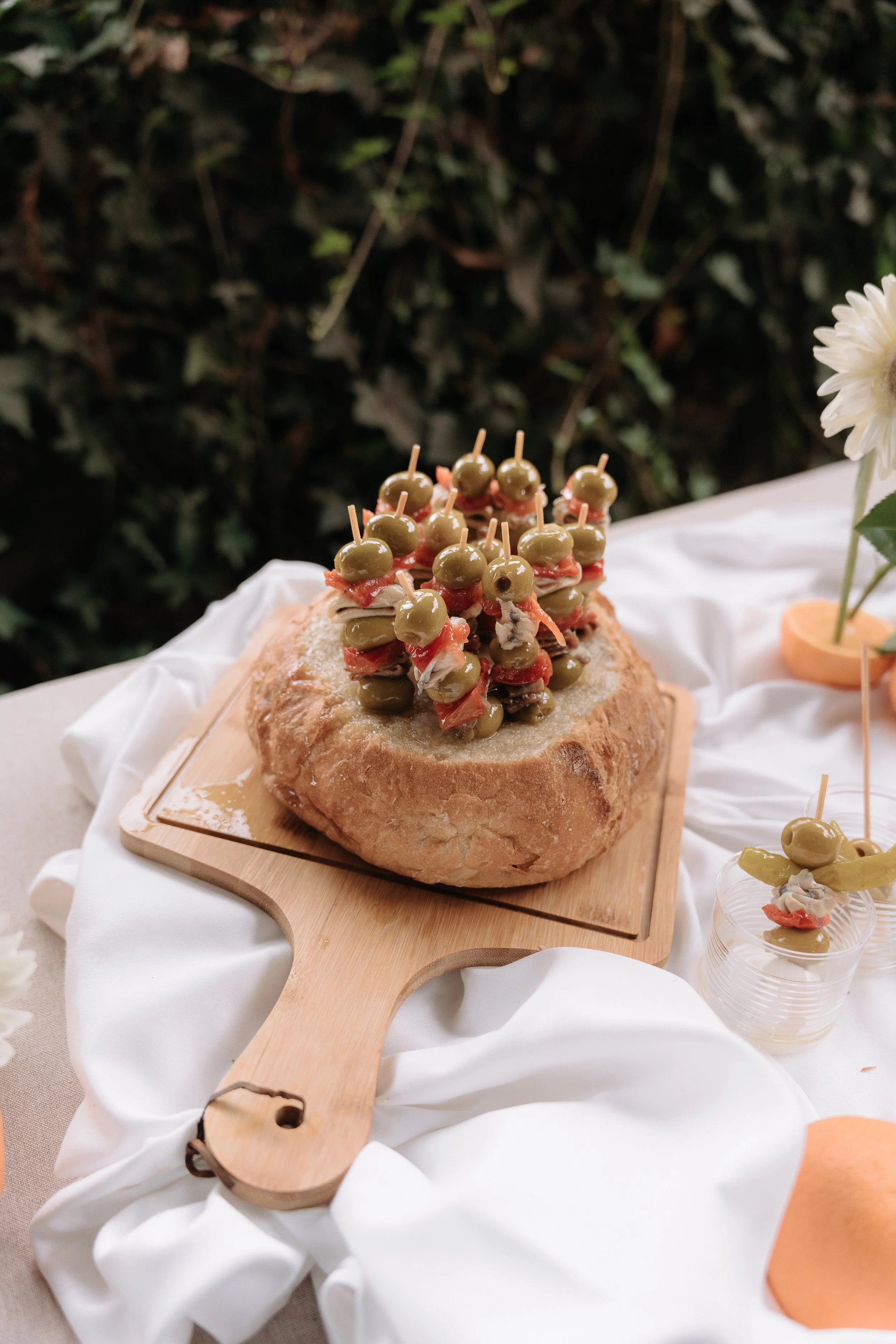 Pan de campo cubierto con aceitunas verdes y jamón, decorado con palillos, sobre una tabla de madera.