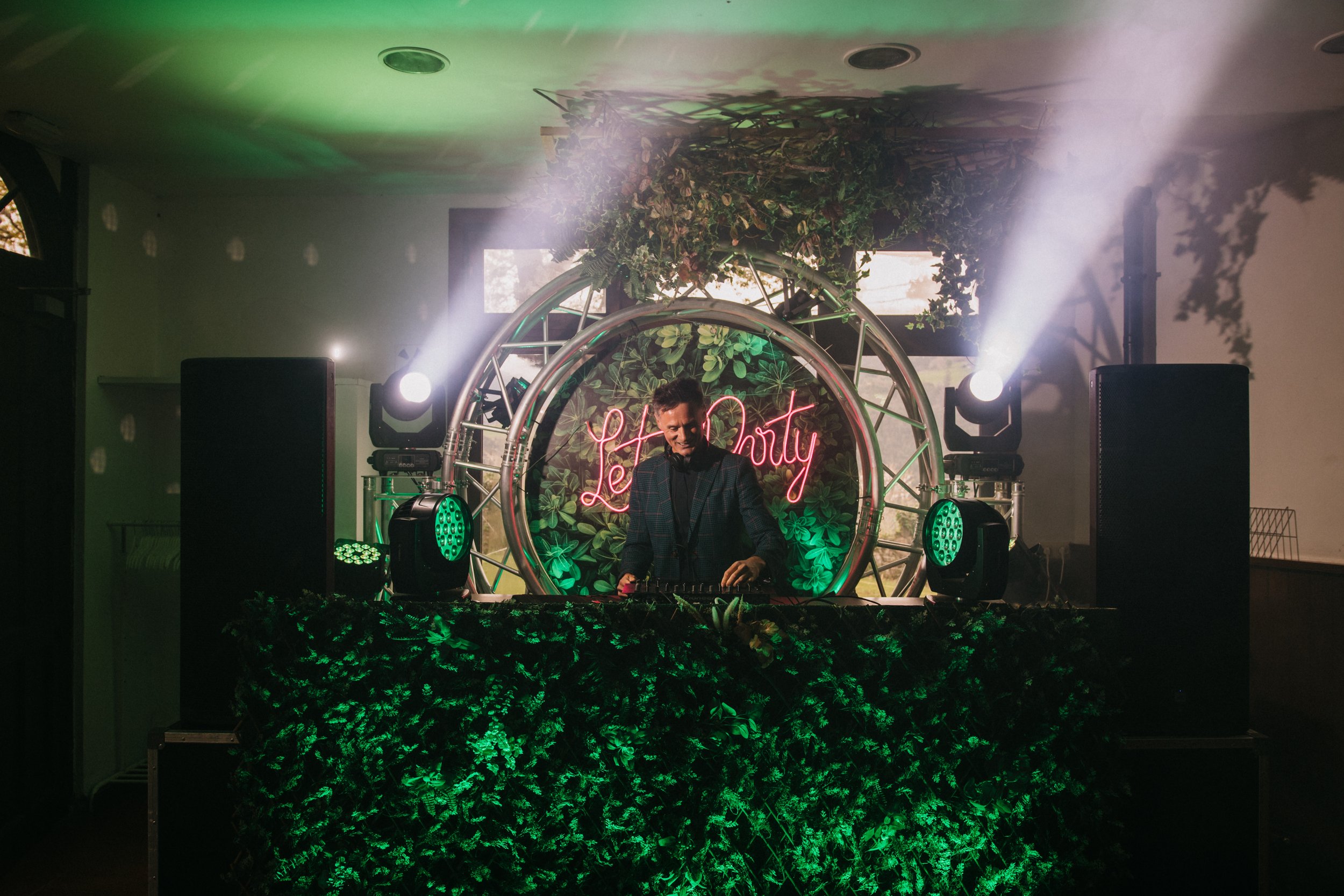 Un DJ tocando en una cabina decorada con muchas plantas y luces, con un letrero de neón que dice 'Let Party' detrás.