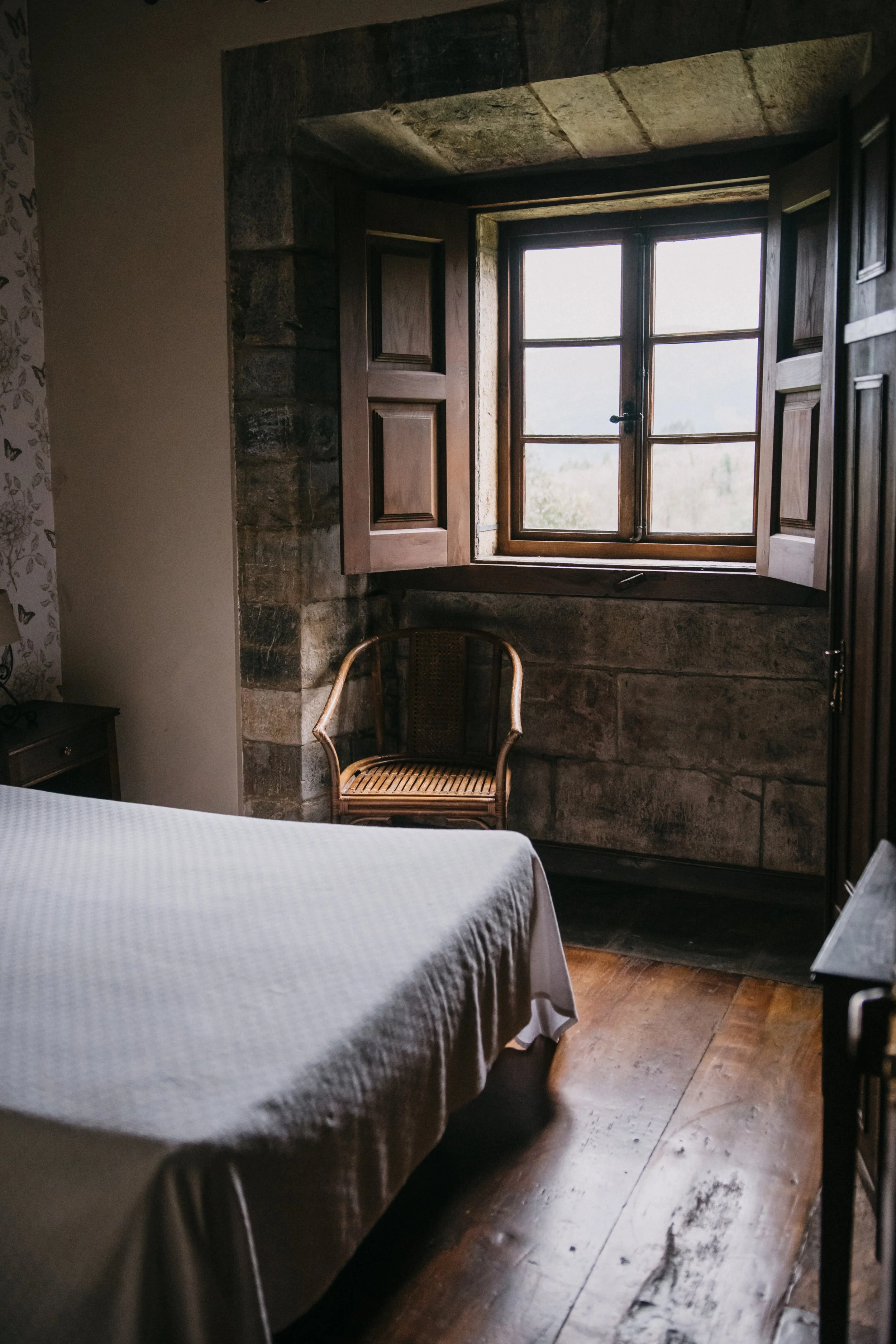 Habitación con cama, ventana de madera con persianas, silla de madera y paredes de piedra.