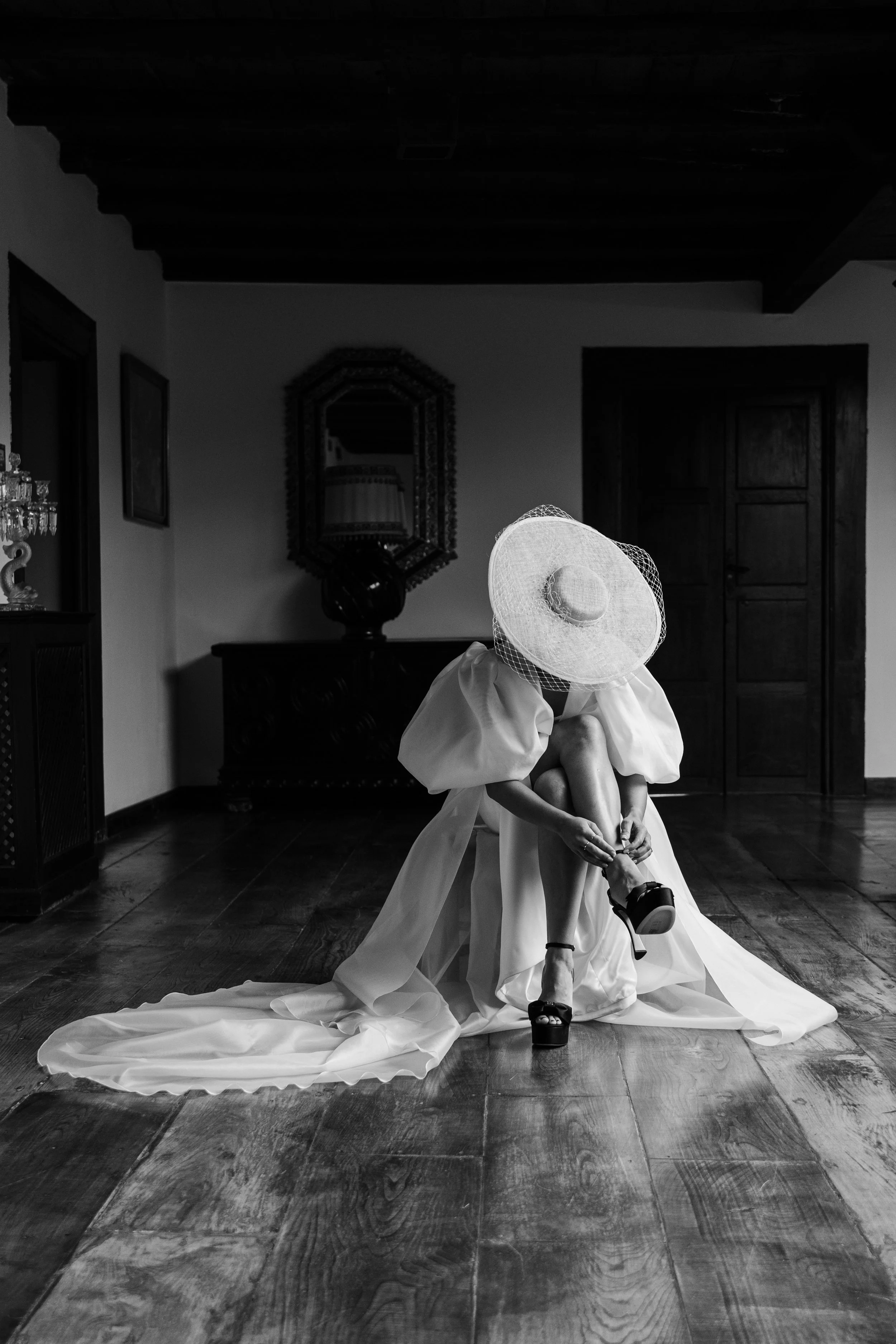 Una mujer con vestido largo y sombrero grande ajustándose una zapatilla en el interior de una habitación.