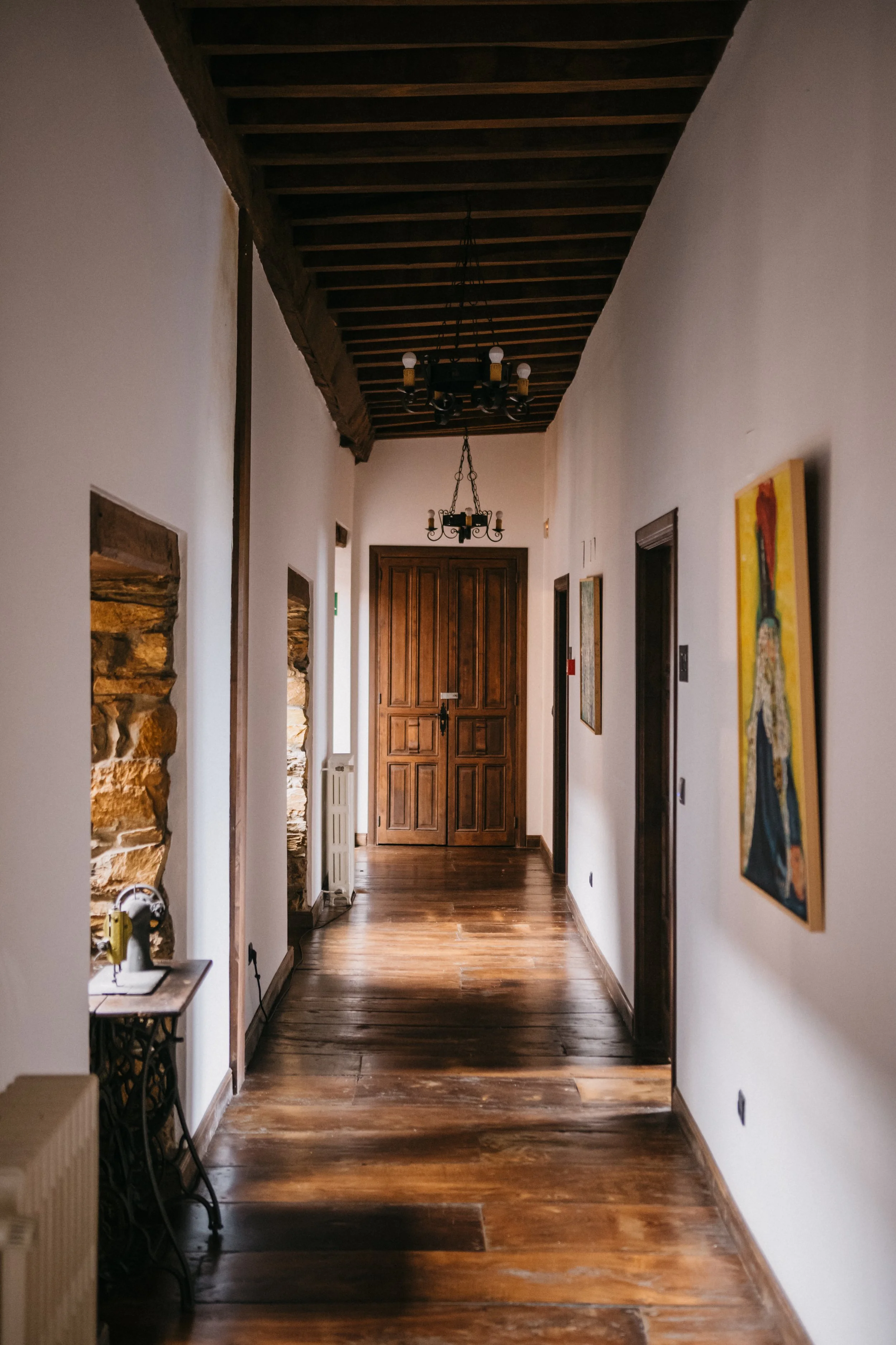 Pasillo interior con paredes blancas, piso de madera, cuadros en las paredes, puertas de madera y candelabros en el techo