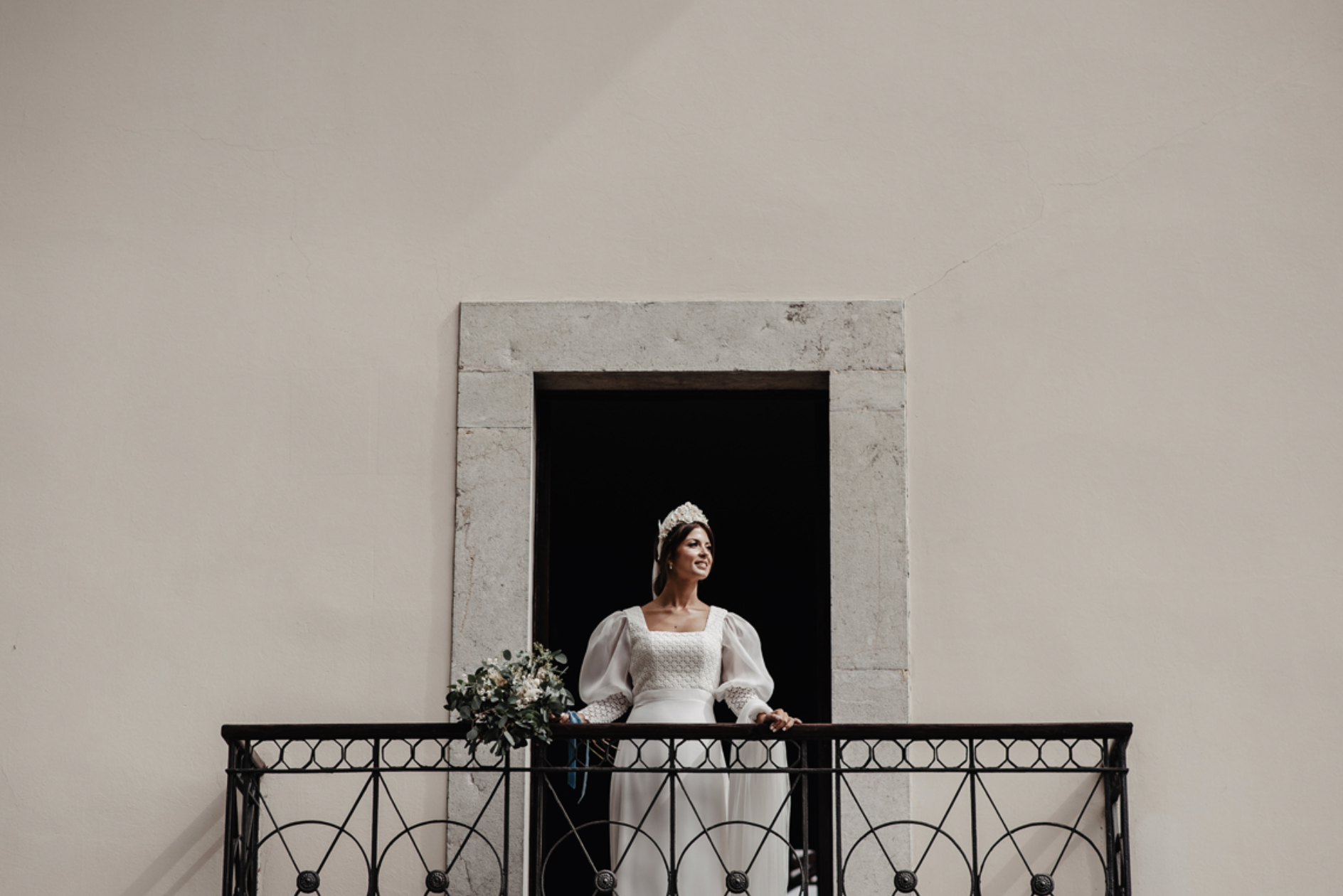 Mujer con vestido de novia en un balcón con barandal de hierro, sosteniendo un ramo de flores, dentro de una estructura de piedra en una pared blanca.