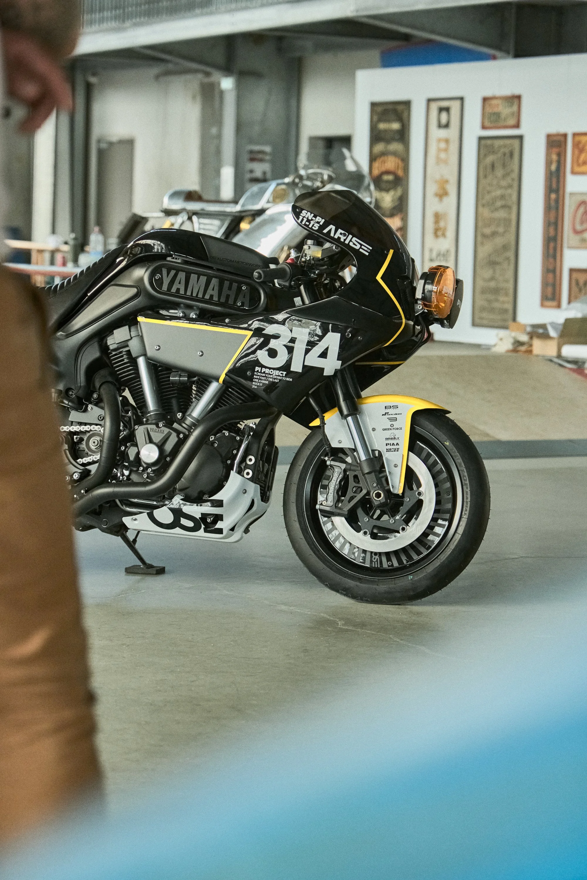 Une moto de course noire avec le numéro 314 et le logo Yamaha, stationnée dans un espace intérieur avec des affiches ou tableaux accrochés sur un mur derrière.