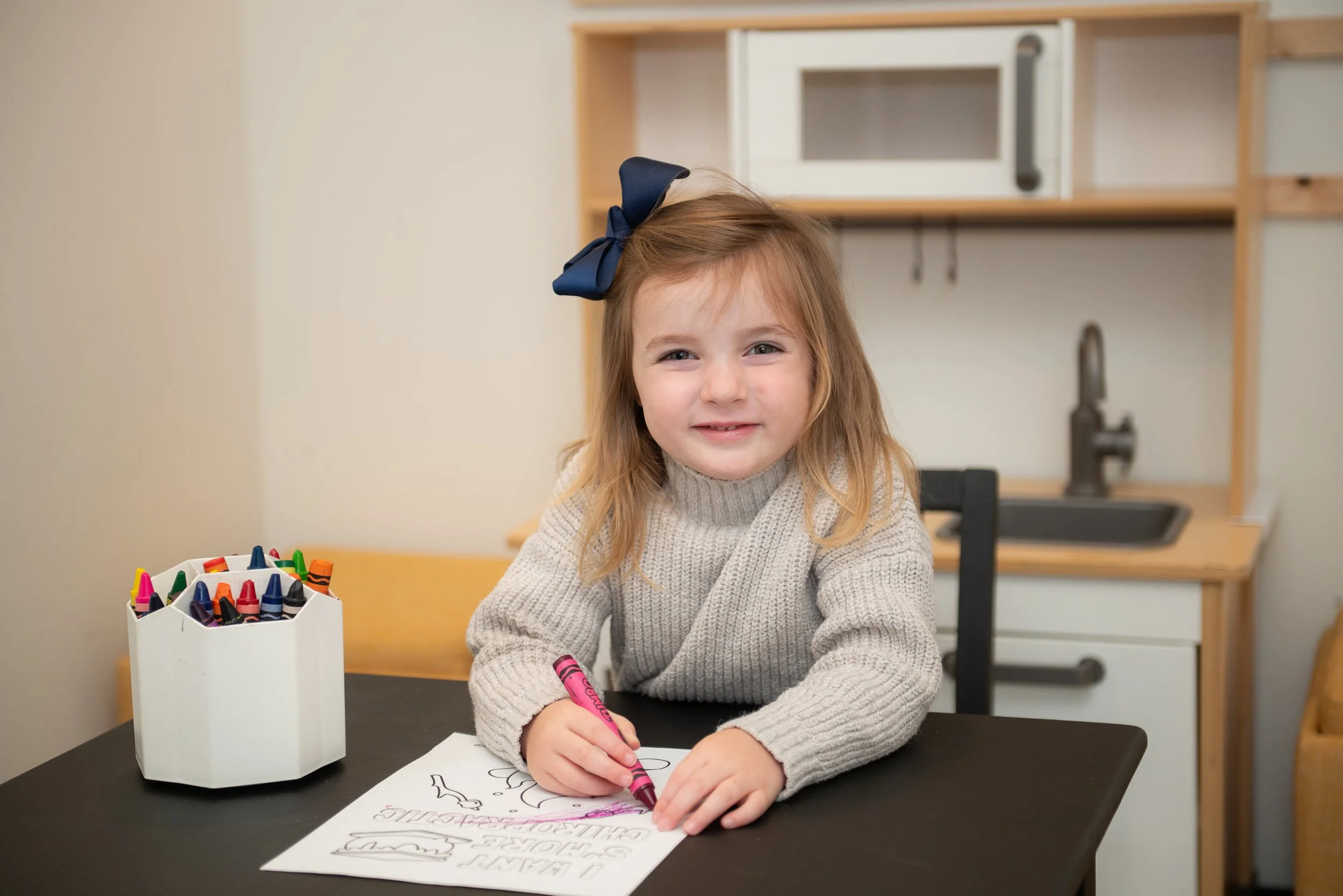 Young child with a blue bow coloring with crayons at a table; a toy kitchen is in the background.
