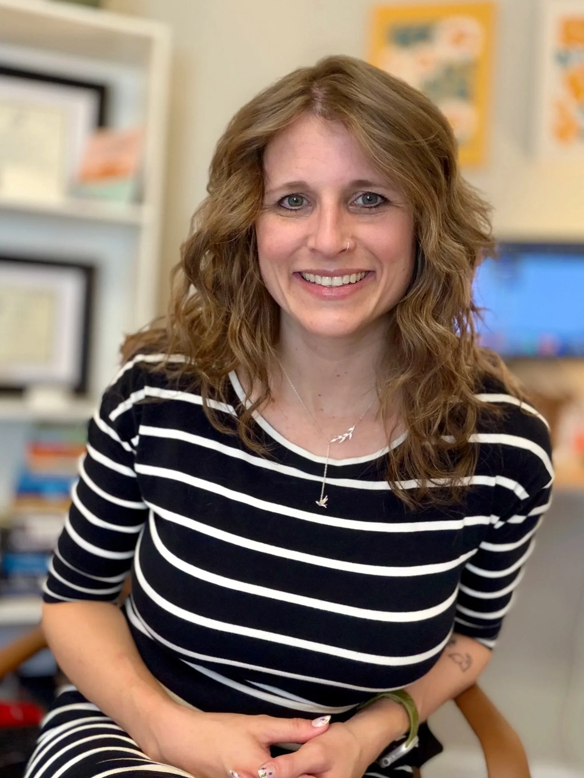 A woman with medium-length, wavy dirty-blonde hair sits smiling in an office chair, hands in her lap. She wears a black-and-white striped long-sleeve top. The background is a warmly lit, modern office with a soft focus.
