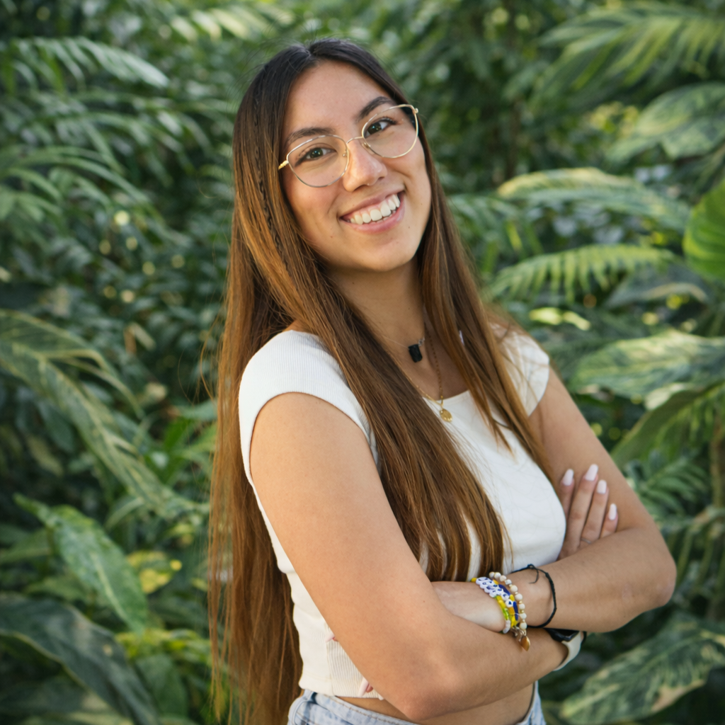 A young woman with long brown hair, wearing glasses, a white sleeveless top, and multiple bracelets, smiling with arms crossed in front of lush green tropical plants.