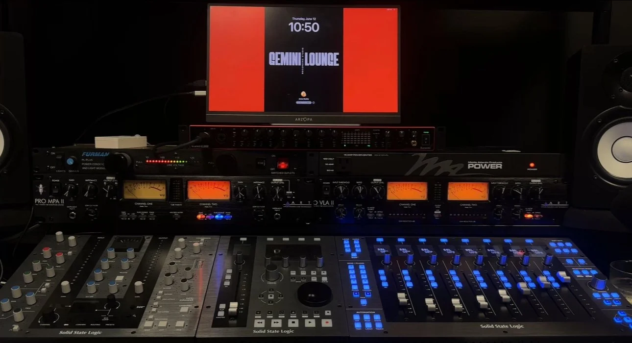 Music production studio setup with a computer displaying digital audio workstation software, studio monitor speakers, a MIDI controller, and audio interface equipment illuminated by purple and orange lights.