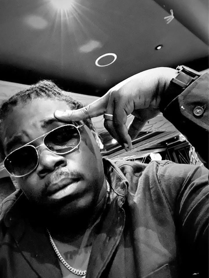 A man wearing sunglasses and a chain necklace, making a gesture with his fingers near his forehead, in a casual setting.