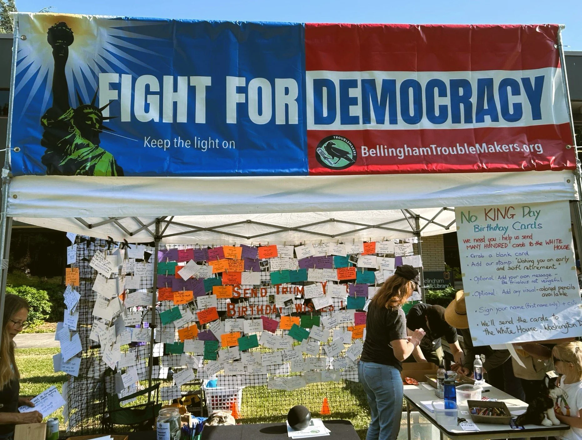 Bellingham Troublemakers hosting a booth during No Kings on June 14, 2025. There is a banner with the text "Fight for Democracy." The backdrop of the booth is netting covered in dozens of colorful "birthday cards" to Trump, written by rally-goers.