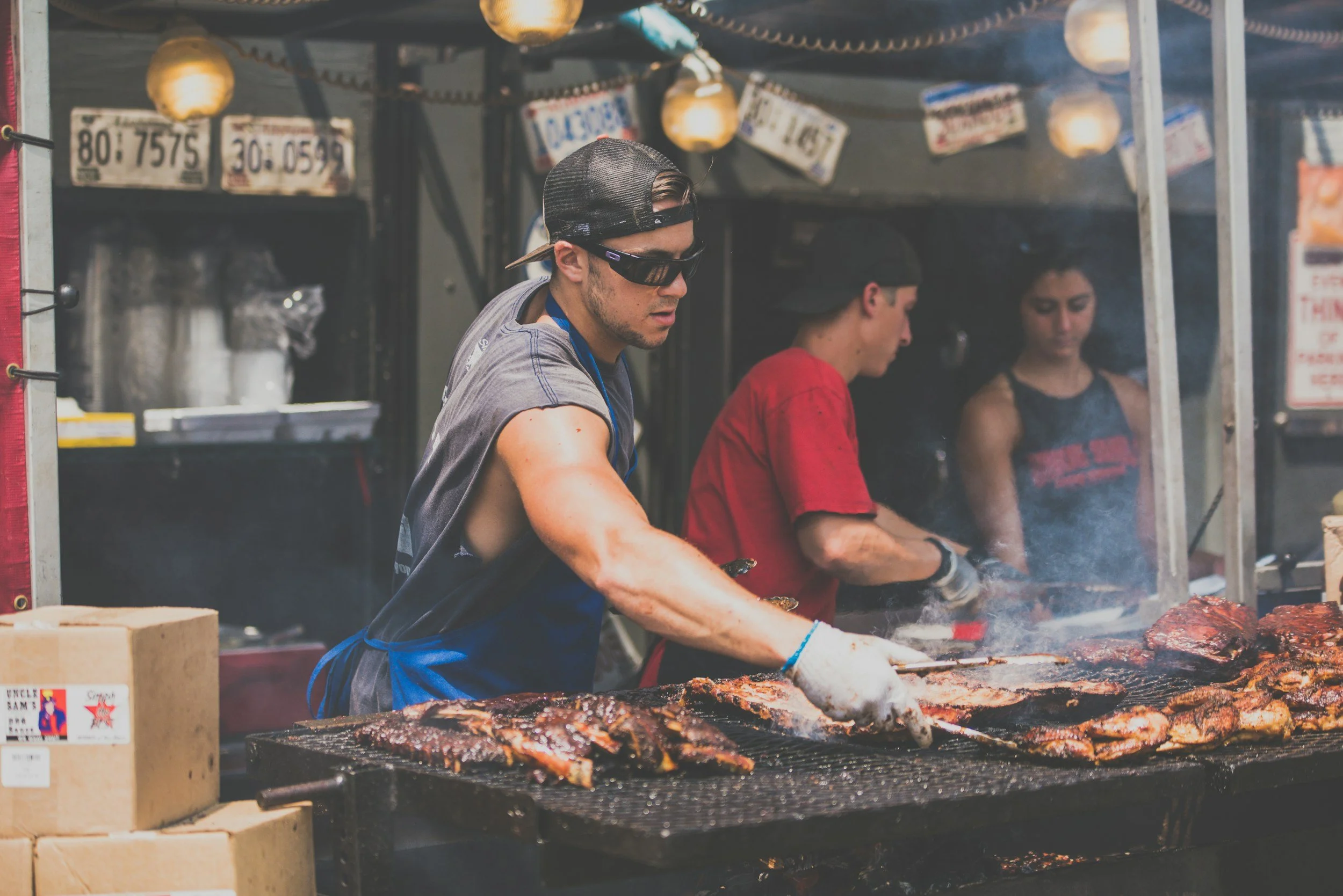 Three people working at a barbecue grill, cooking meat with smoke and flames, at a food stall with hanging lanterns and signs in the background.