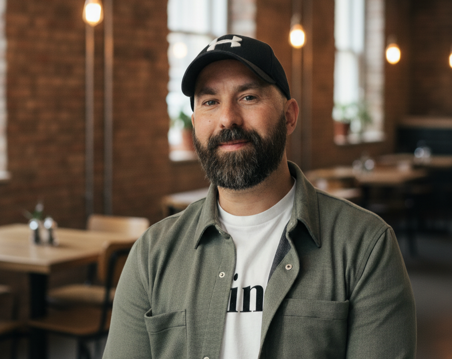 A bearded man wearing a black Under Armour cap and gray shirt, sitting in a cozy restaurant with exposed brick walls, pendant lights, and wooden tables and chairs.