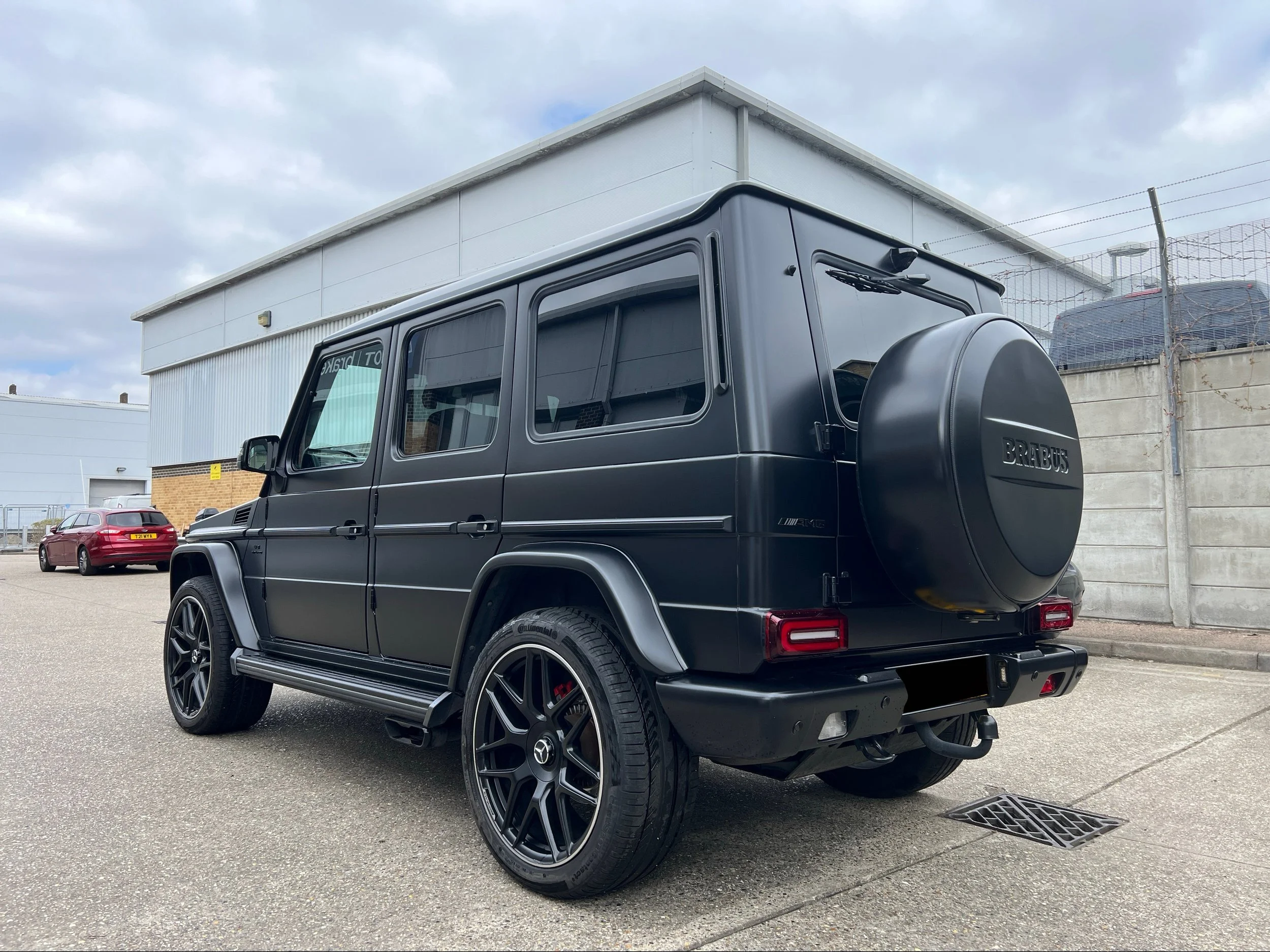 A black Mercedes-Benz G-Class SUV parked in an industrial lot. The vehicle has custom black rims, a spare tire cover labeled 'BRABUS,' tinted windows, and is positioned in front of a building with a metal fence and cloudy sky.