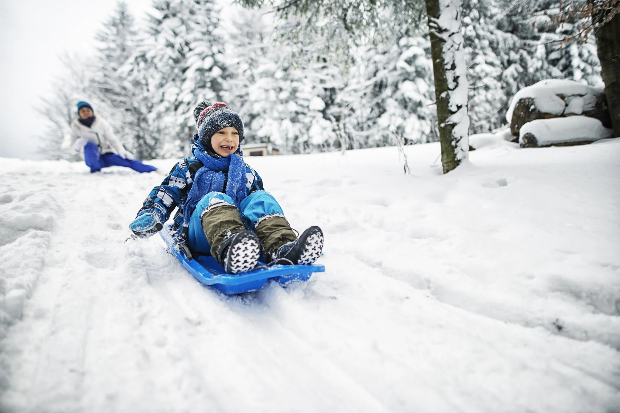 Children playing in the snow with one child sledding and another child in the background on a snowy hillside surrounded by snow-covered trees.