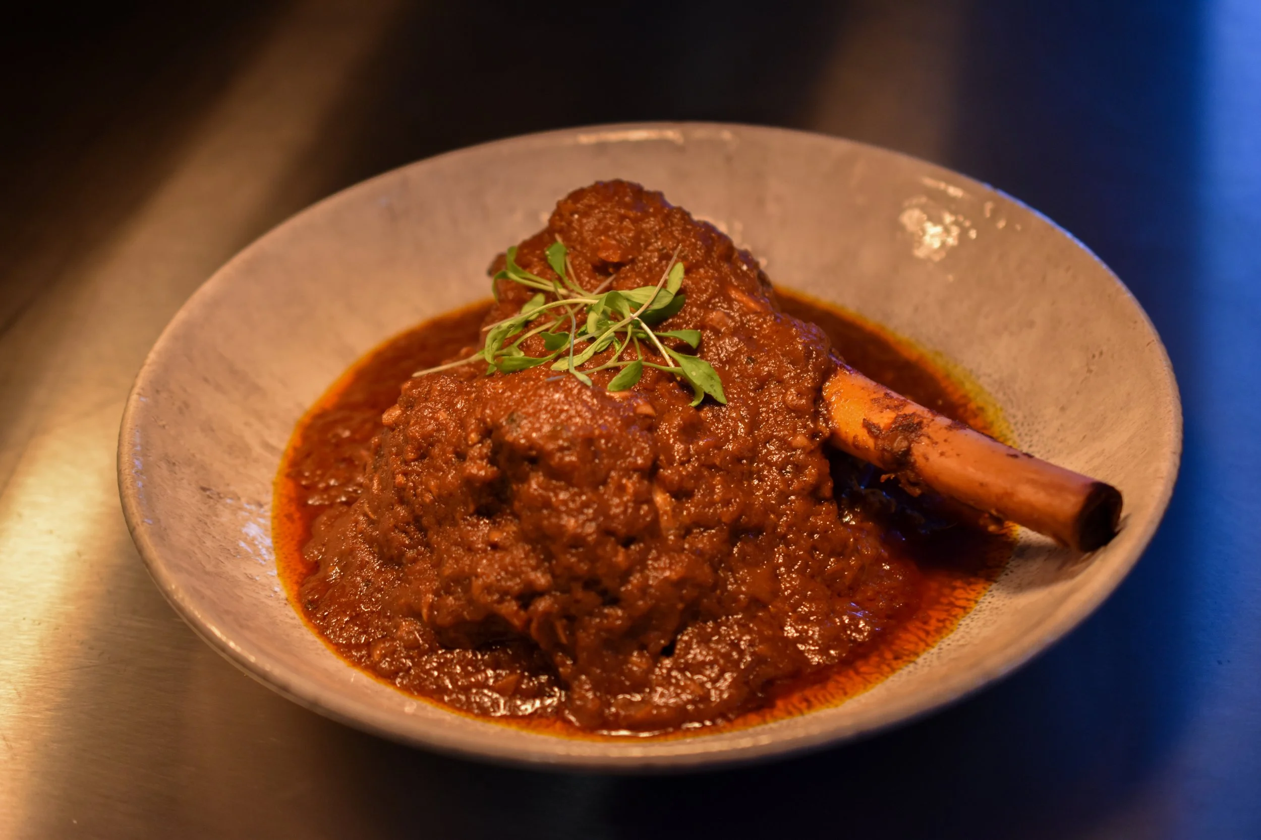 A dish of spicy meat curry with a bone-in portion, garnished with fresh green microgreens in a beige bowl.