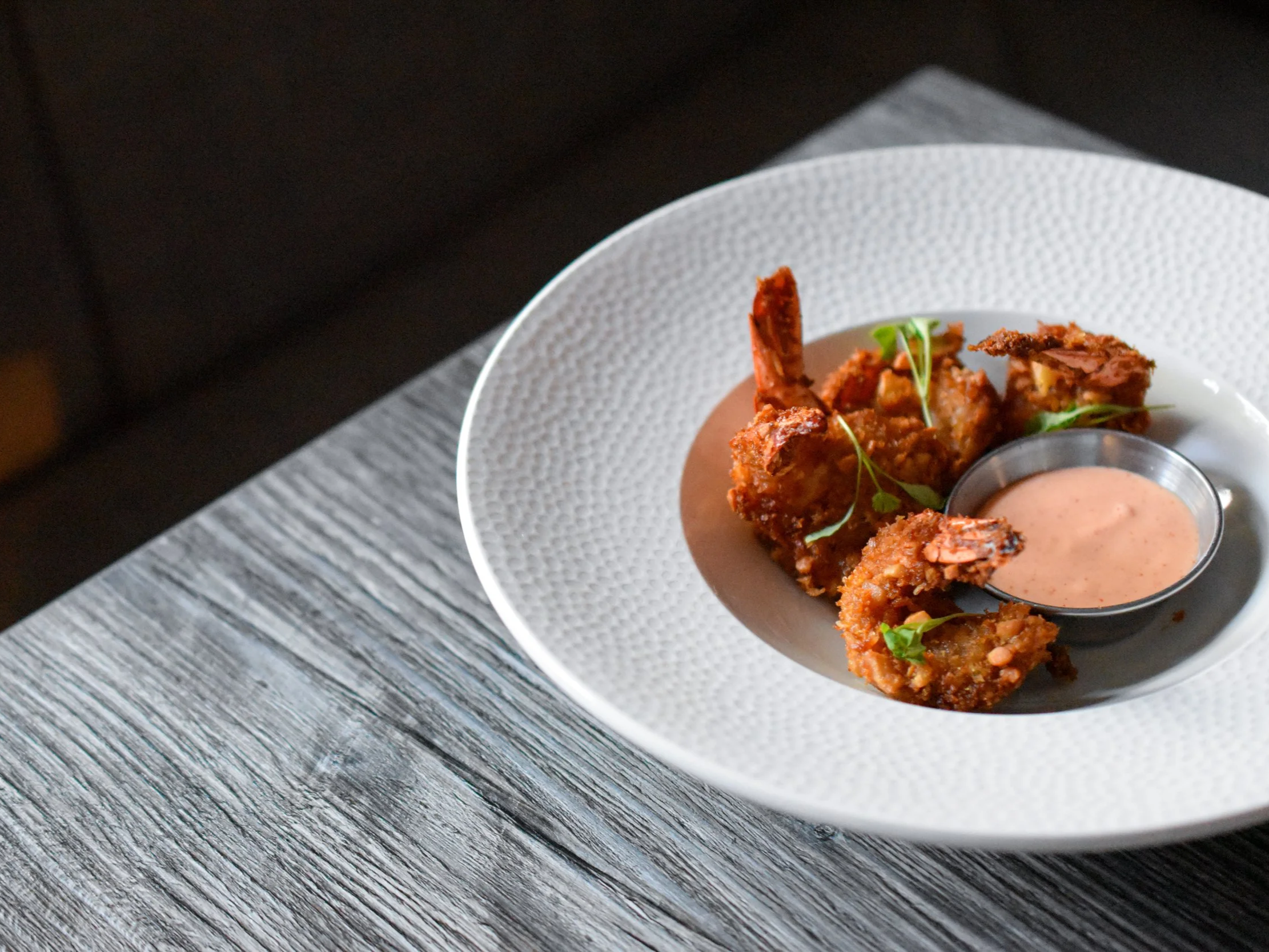 Fried chicken pieces on a white, textured plate with a small bowl of pink dipping sauce and green garnish, on a gray wooden surface.
