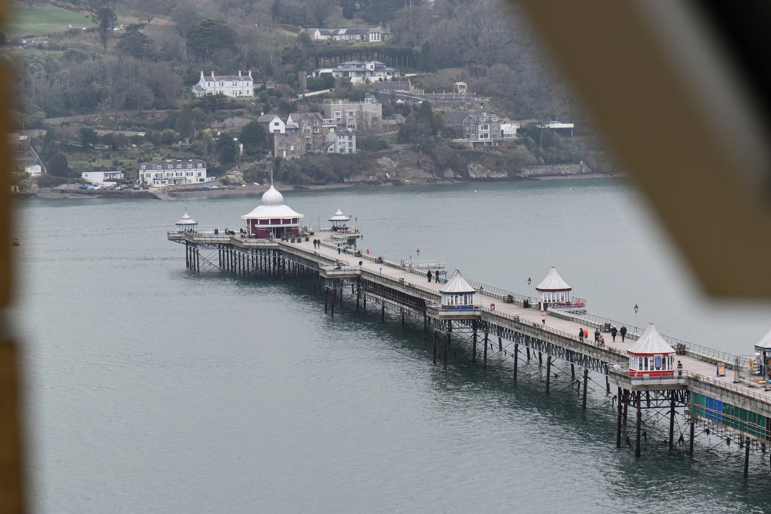 A pier extending into a body of water, with small pavilions at intervals, leading towards a shoreline with houses and trees on a hillside.