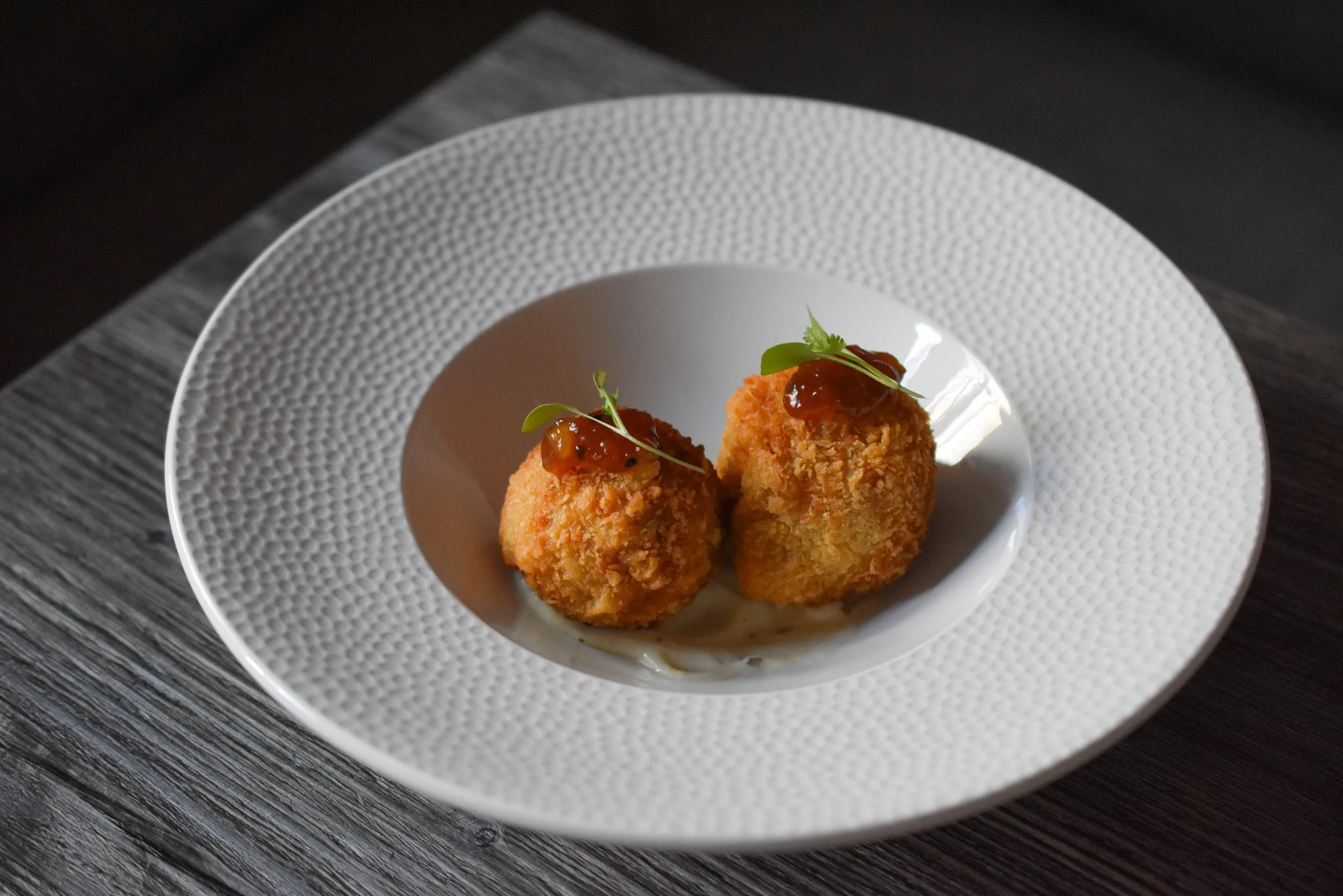 Two fried appetizer balls with sauce and garnished with microgreens on a textured white plate, placed on a dark wooden surface.