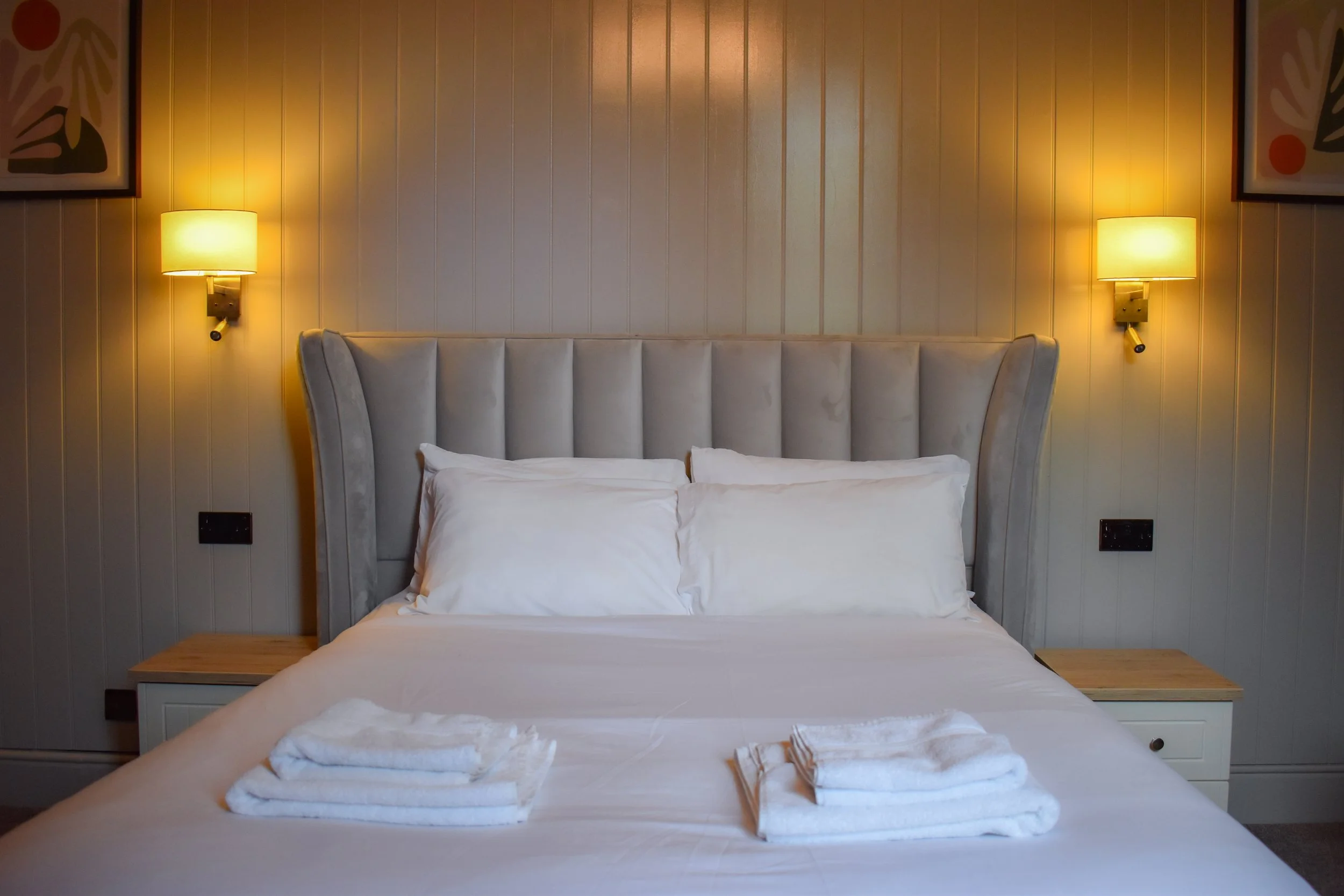A neatly made bed with white pillows and a folded towel on top, flanked by two wall-mounted lamps on a beige paneled wall with framed artwork.