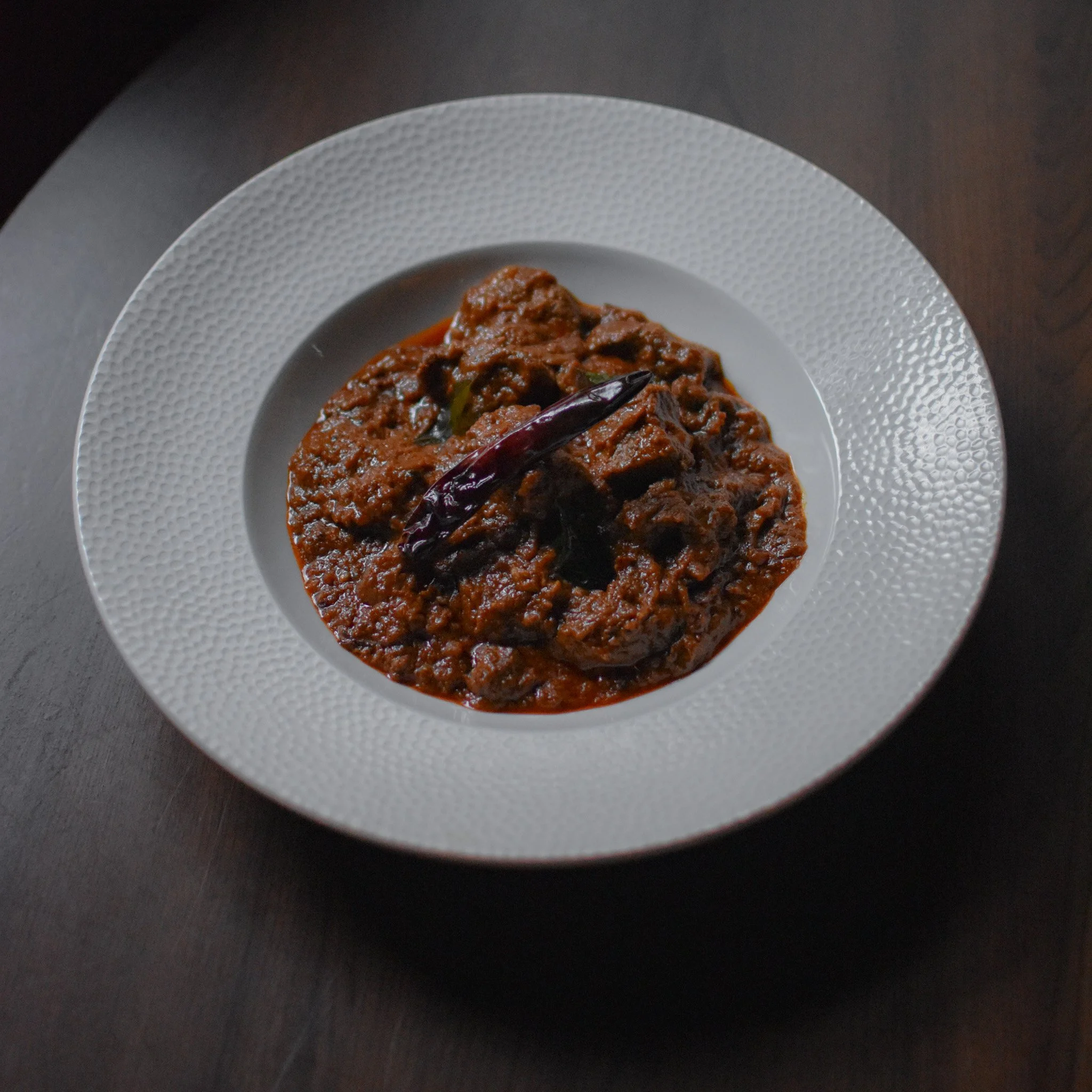 A white textured plate with a beef stew garnished with dried chili peppers placed on a dark surface.