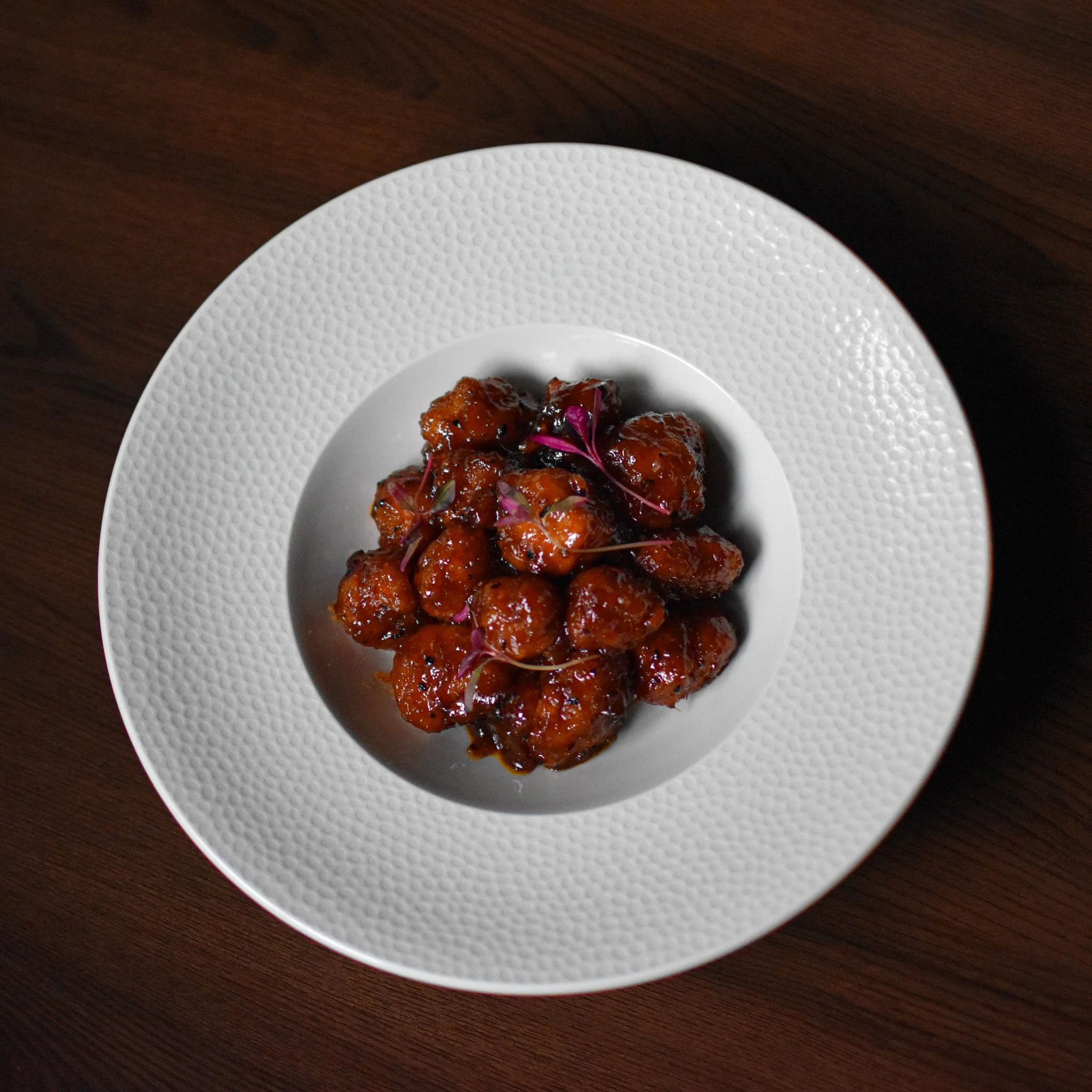 A white textured plate with glazed pieces of food, garnished with pink edible flowers, on a dark wooden surface.