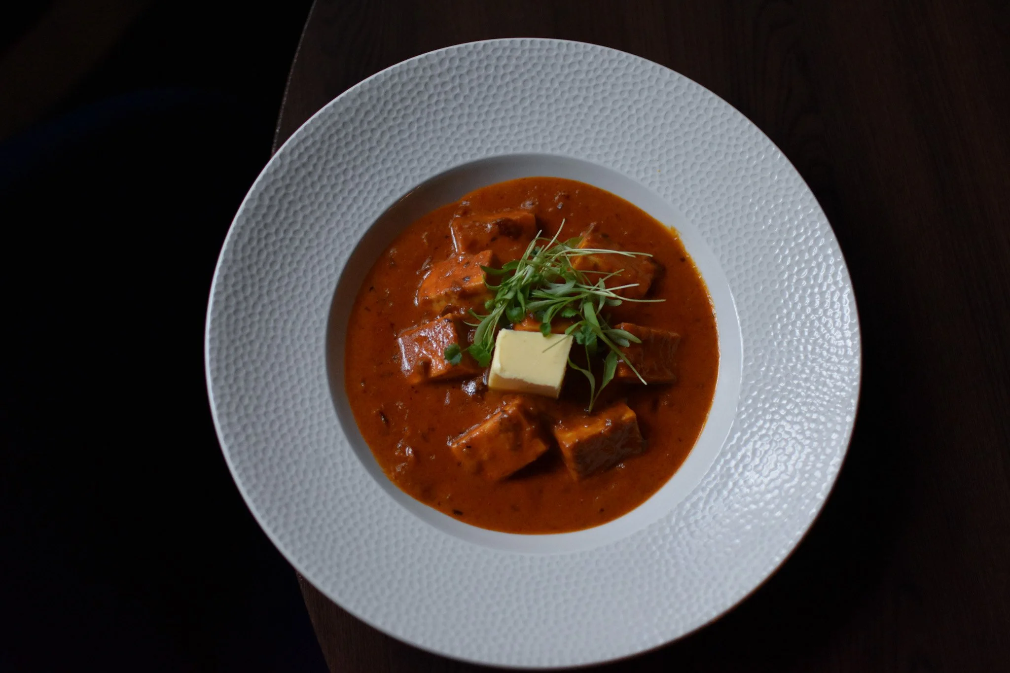 A bowl of red stew with chunks of meat, garnished with microgreens and a pat of butter, on a white textured plate.