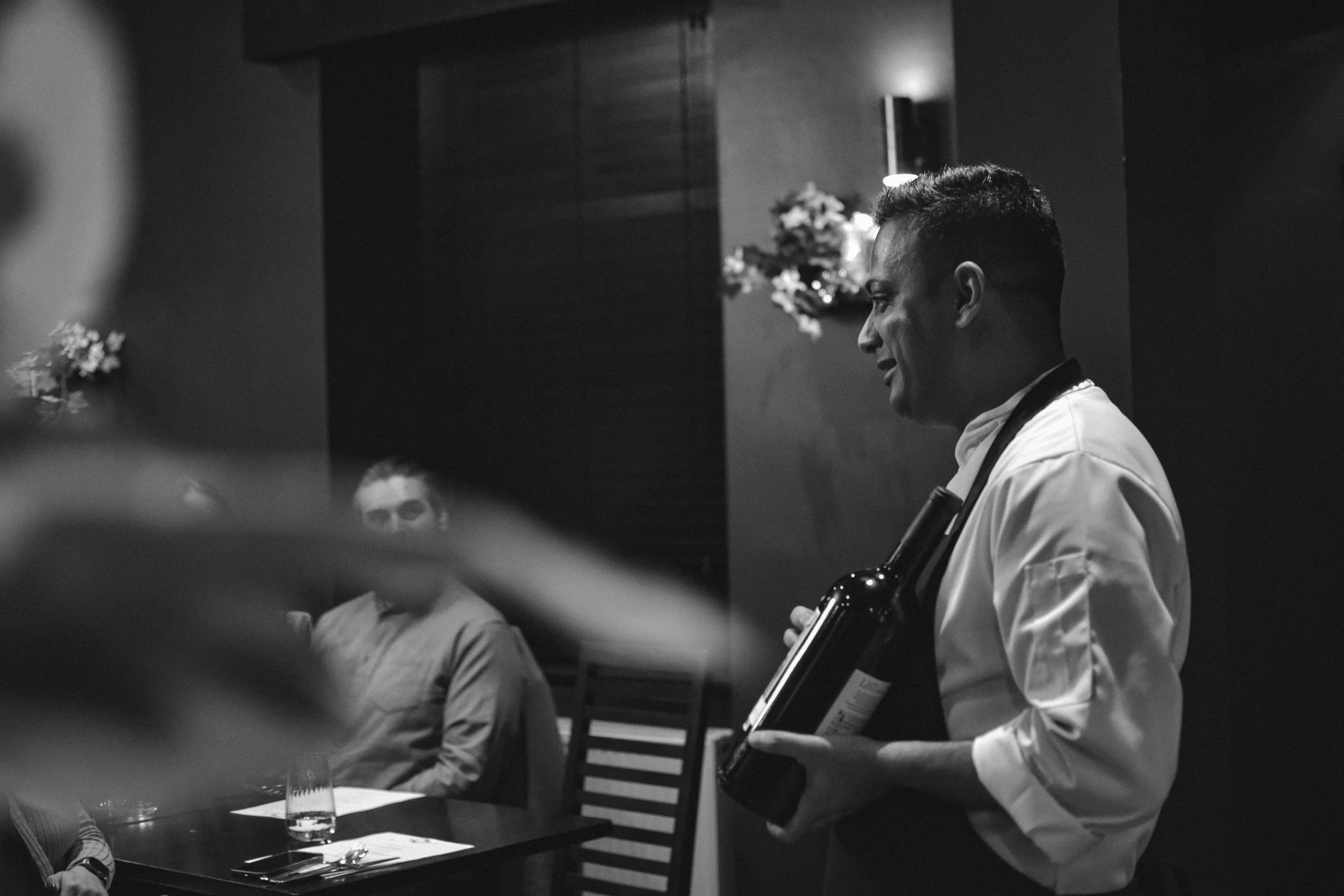 A man in a white shirt and apron, possibly a waiter or chef, holding a wine bottle with a glass of water and a glass of wine on the table. Several people sitting at a table in a restaurant setting, with flowers on the wall behind them.