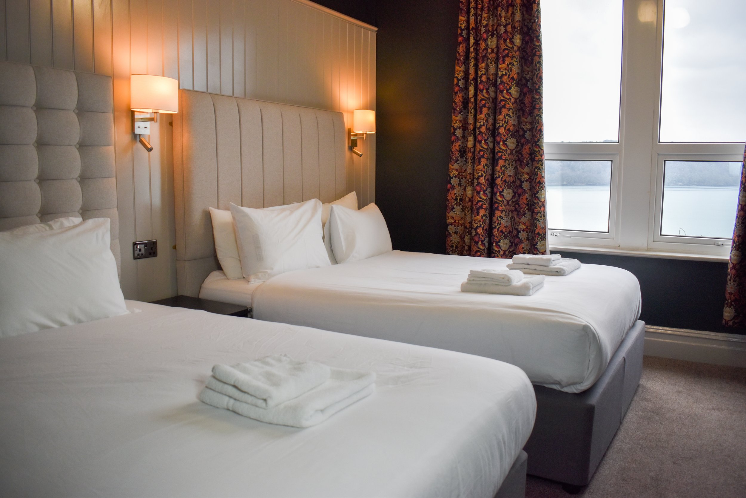 Hotel room with two neatly made beds, each with white linens and pillows, topped with folded towels. The room features a large window with patterned curtains, beige padded headboards, and wall-mounted lamps on a wood-paneled wall.