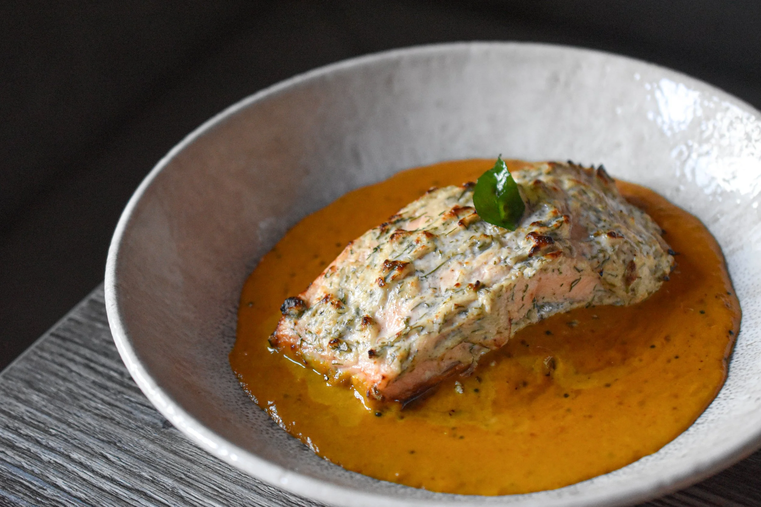 A bowl of cooked salmon topped with herb crust, sitting in a creamy sauce with a green garnish on top.