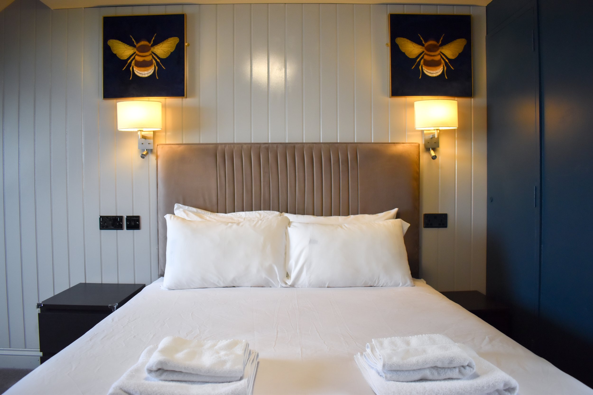 A neatly made bed with white linens and pillows, two towels placed at the foot of the bed, with wall-mounted lamps and two bee-themed paintings above the headboard in a bedroom.