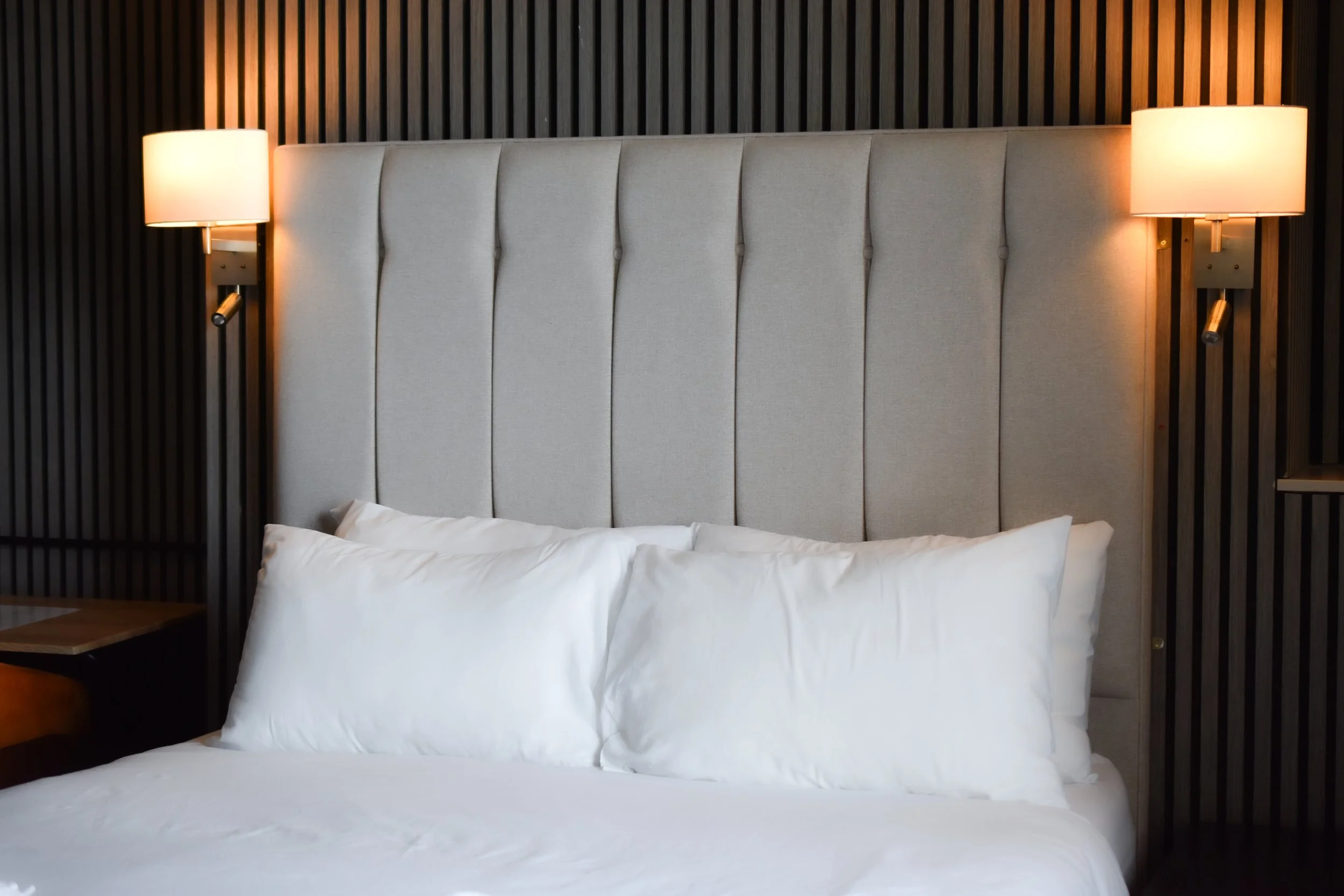 A neatly made bed with white pillows and bedding, a beige upholstered headboard, flanked by wall-mounted lamps with warm lighting, against a dark wooden panel wall.