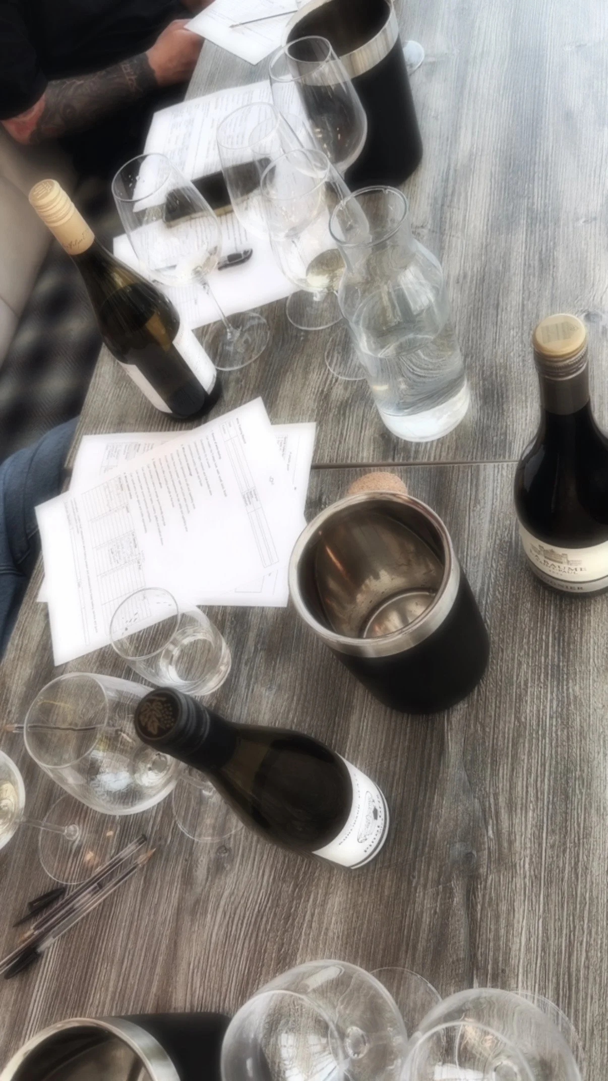 A table set with wine bottles, empty wine glasses, a water pitcher, papers, and pens, with some people seated around.