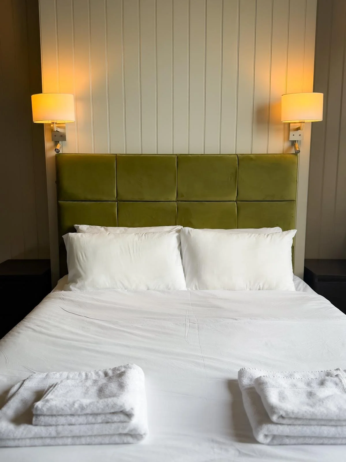 A neatly made bed with white sheets and two pillows, featuring a green upholstered headboard, in a hotel room with wall-mounted lamps on either side.