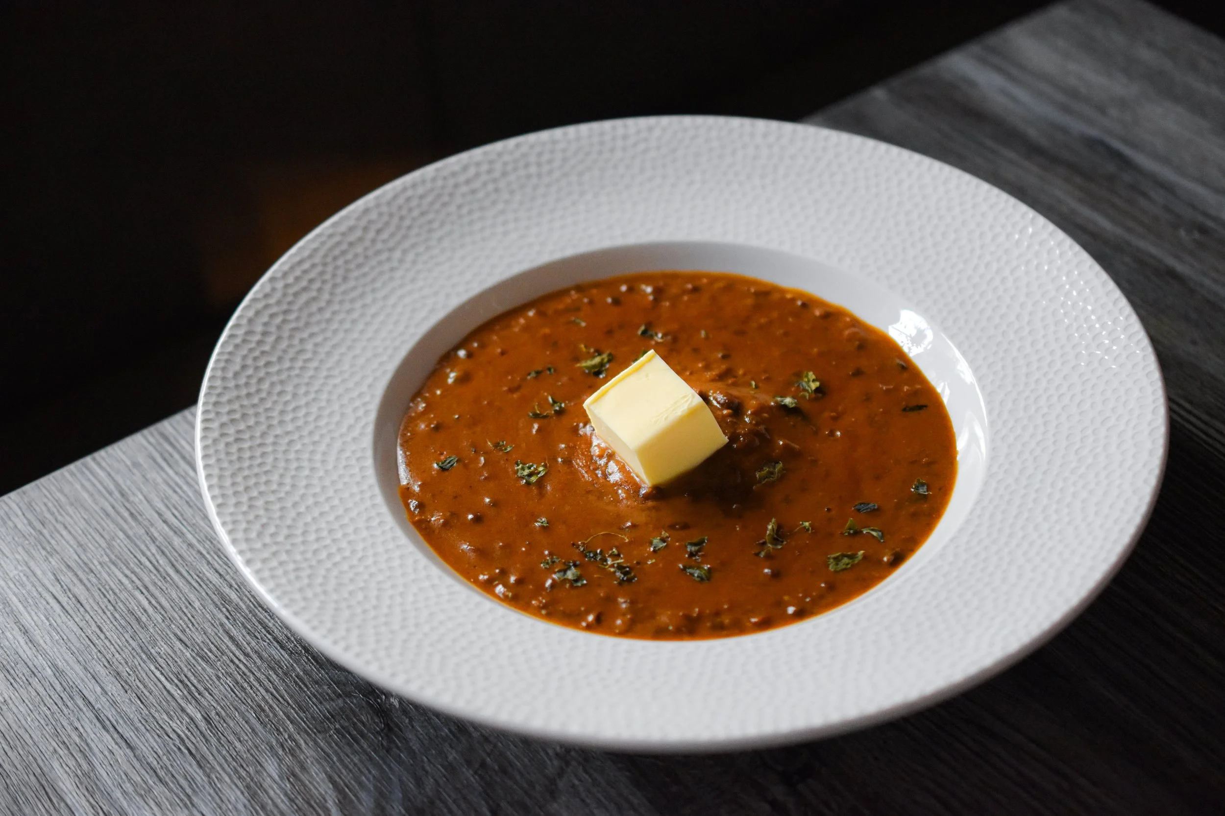A white textured bowl filled with tomato-based soup garnished with herbs and a pat of butter on top, placed on a wooden table.