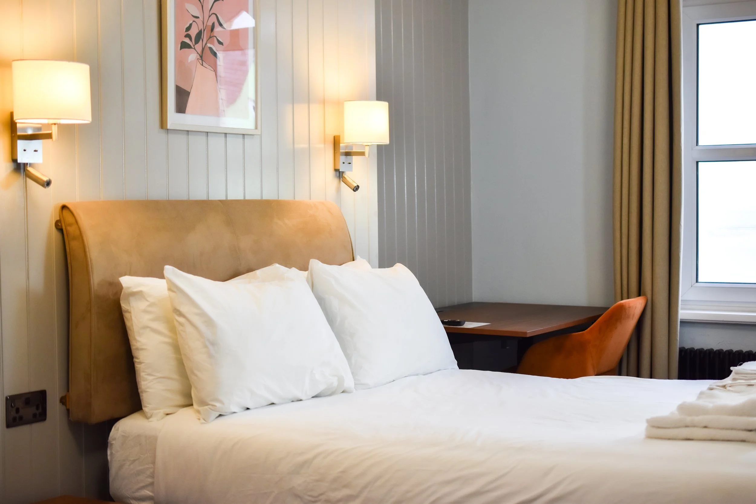A neatly made bed with multiple white pillows, a beige headboard, and white linens in a modern bedroom. Two wall-mounted lamps are on either side of the bed, and there is a framed abstract art print above the headboard. A window with beige curtains is on the right side of the image.