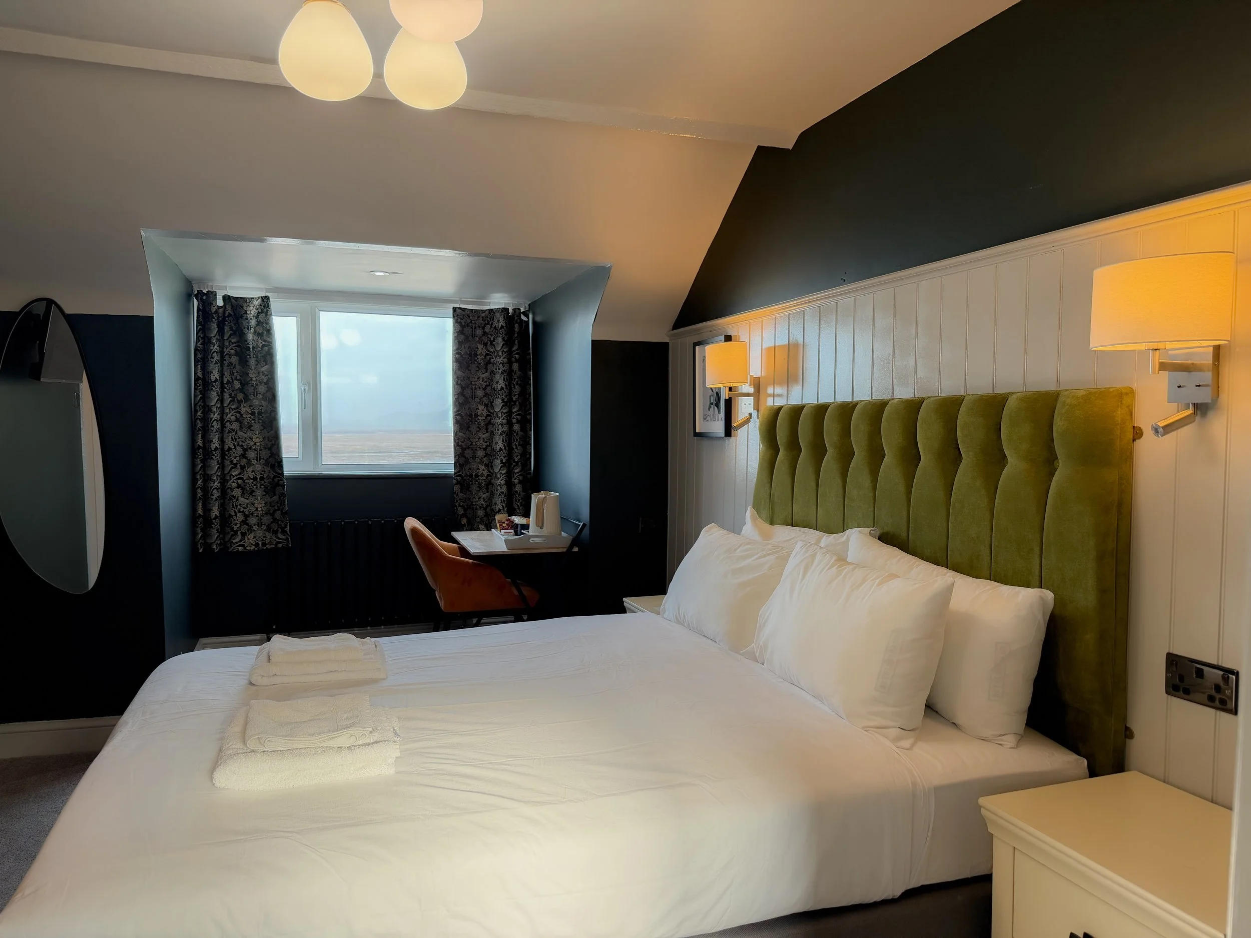 A hotel room with a white bed, green tufted headboard, white linens, and folded white towels on the bed. There are wall-mounted lamps, a seating chair, and a window with dark floral curtains showing an outdoor view.