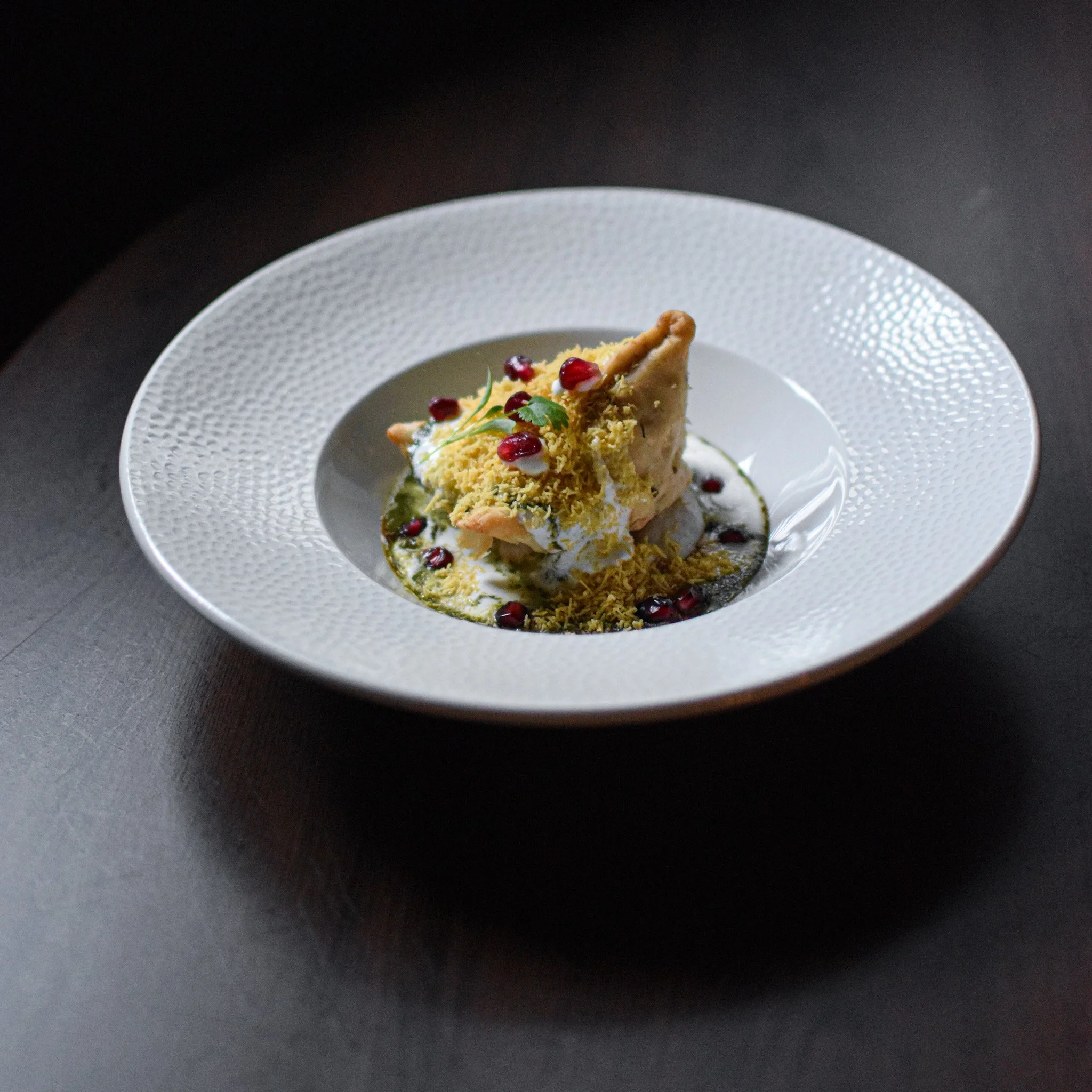 A white textured bowl holds a slice of cake topped with pomegranate seeds and herbs, served on a dark surface.