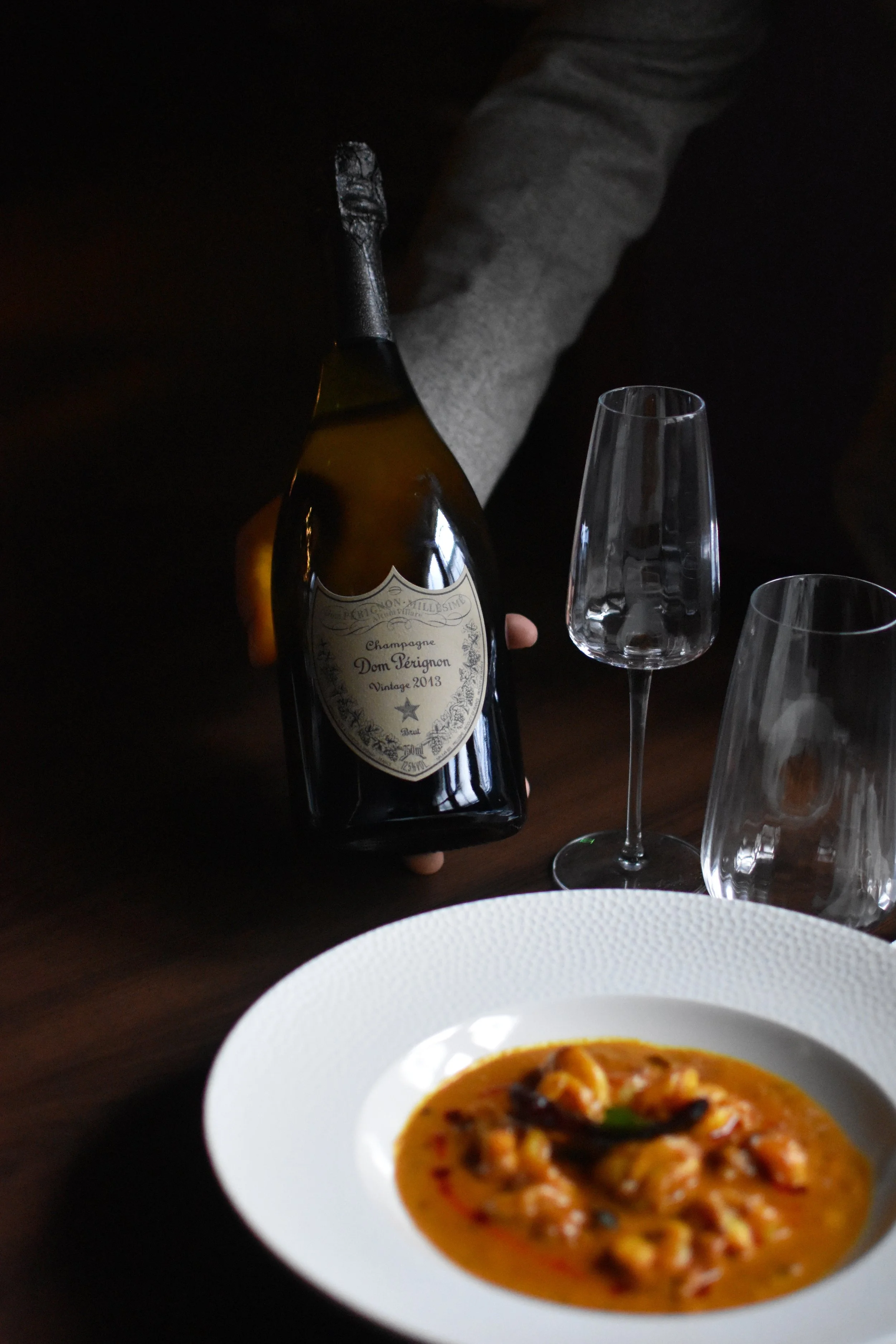 A person holding a bottle of Dom Pérignon vintage 2013 champagne, with two empty champagne flutes nearby. In the foreground, there is a white bowl of soup with vegetables.