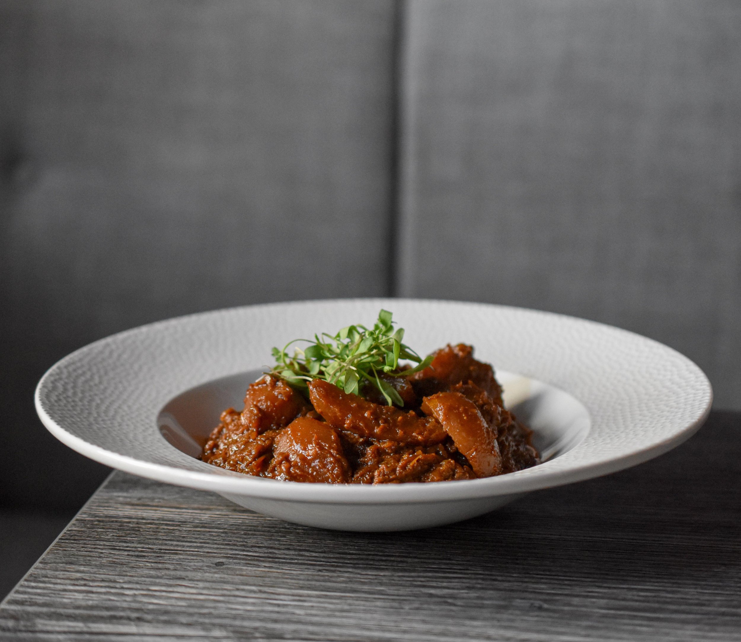 A white bowl of thick, brown stew garnished with green microgreens, placed on a dark wooden surface against a black background.