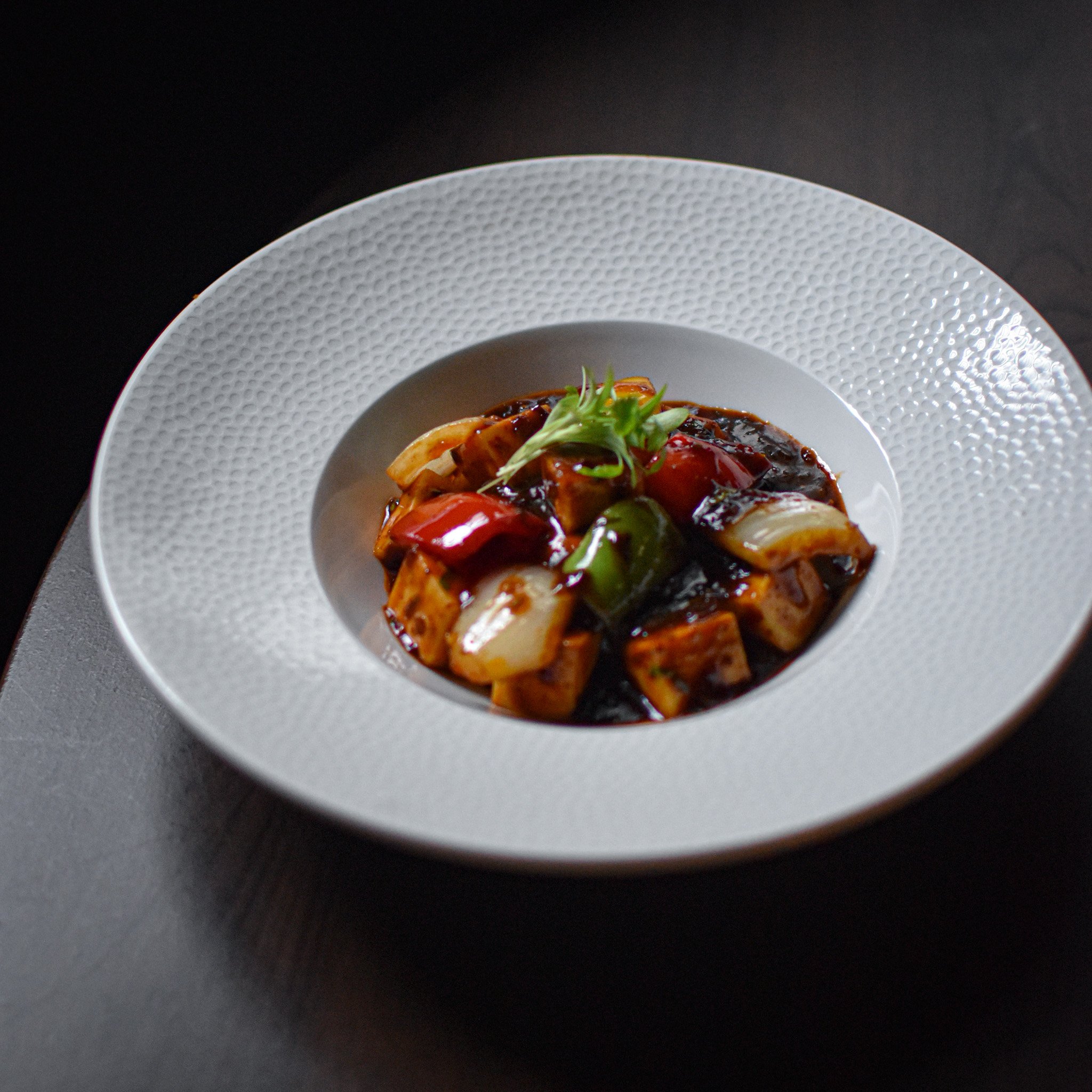 Close-up of a white textured plate with a gourmet dish topped with greens on a dark surface.