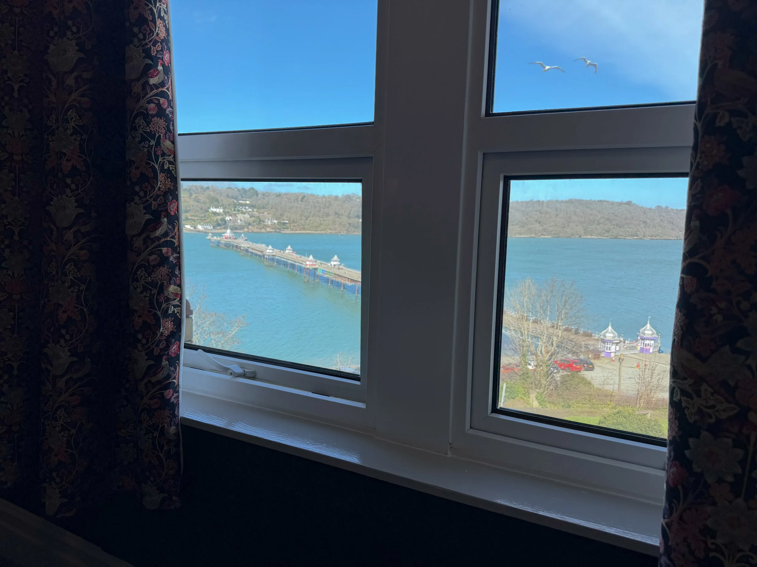 View through a window showing a clear blue sky, a distant horizon with hills, a body of water, and a pier with small buildings. Dark floral curtains frame the window.